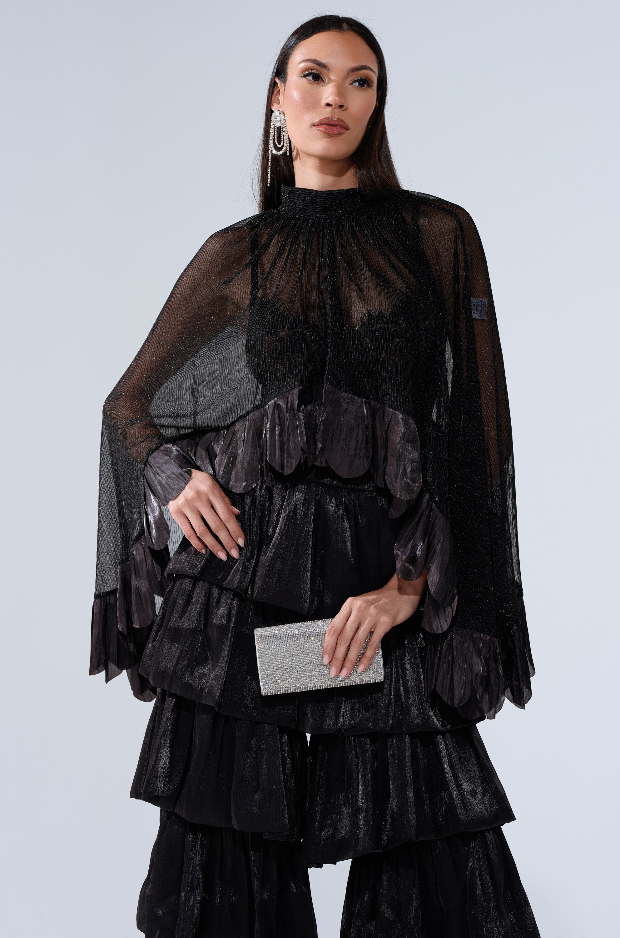 A woman in the BRIGHT LIFE BLOUSE, featuring sheer fabric and wide sleeves, pairs it with a tiered black ruffle skirt and a silver glitter clutch. She wears statement earrings and stands confidently against a light gray background.