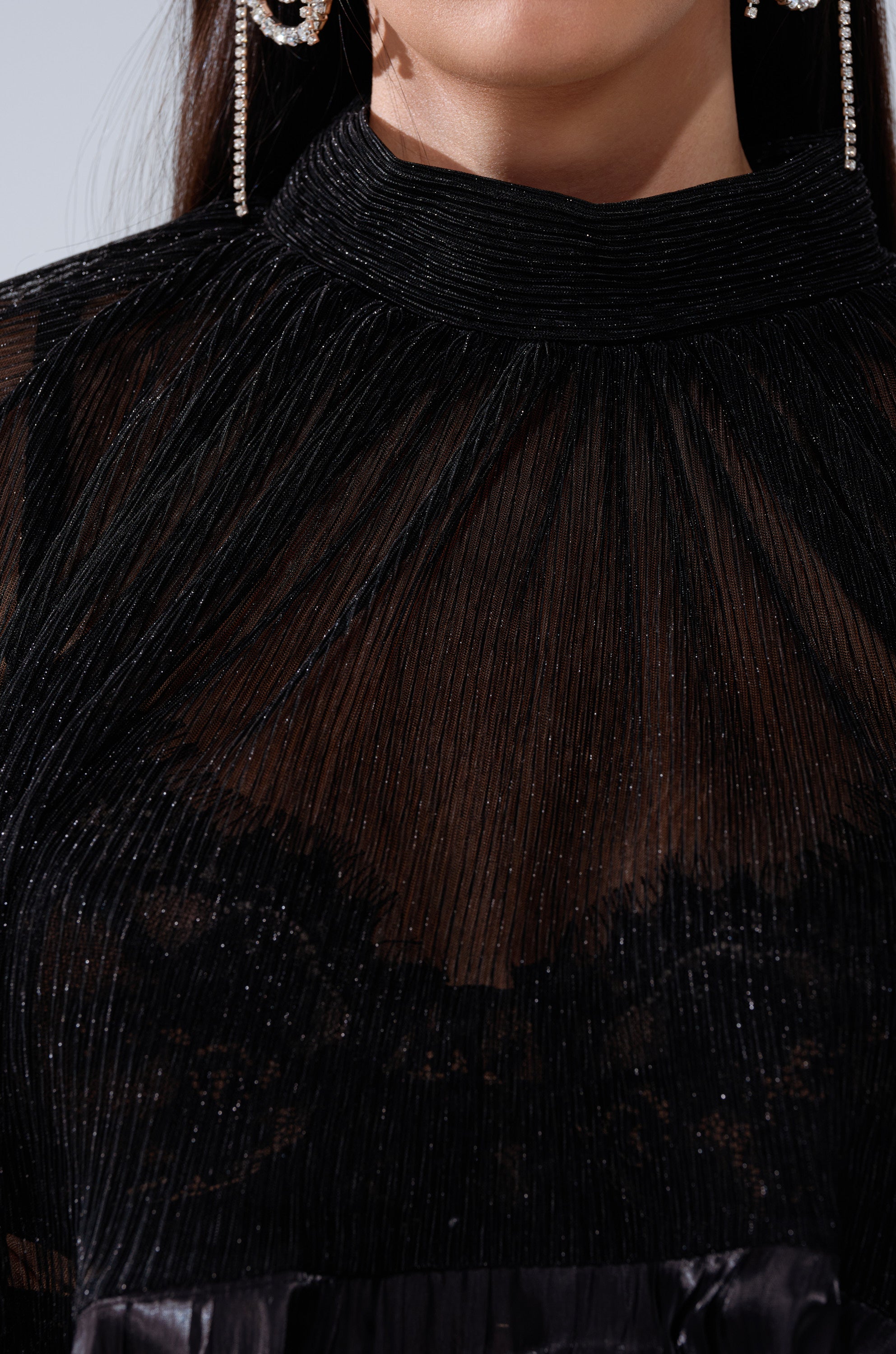 Close-up of a woman wearing the BRIGHT LIFE BLOUSE—a sheer plissé top with a high neck and subtle sparkle, revealing an opaque black layer underneath. She pairs it with large, dangling earrings.