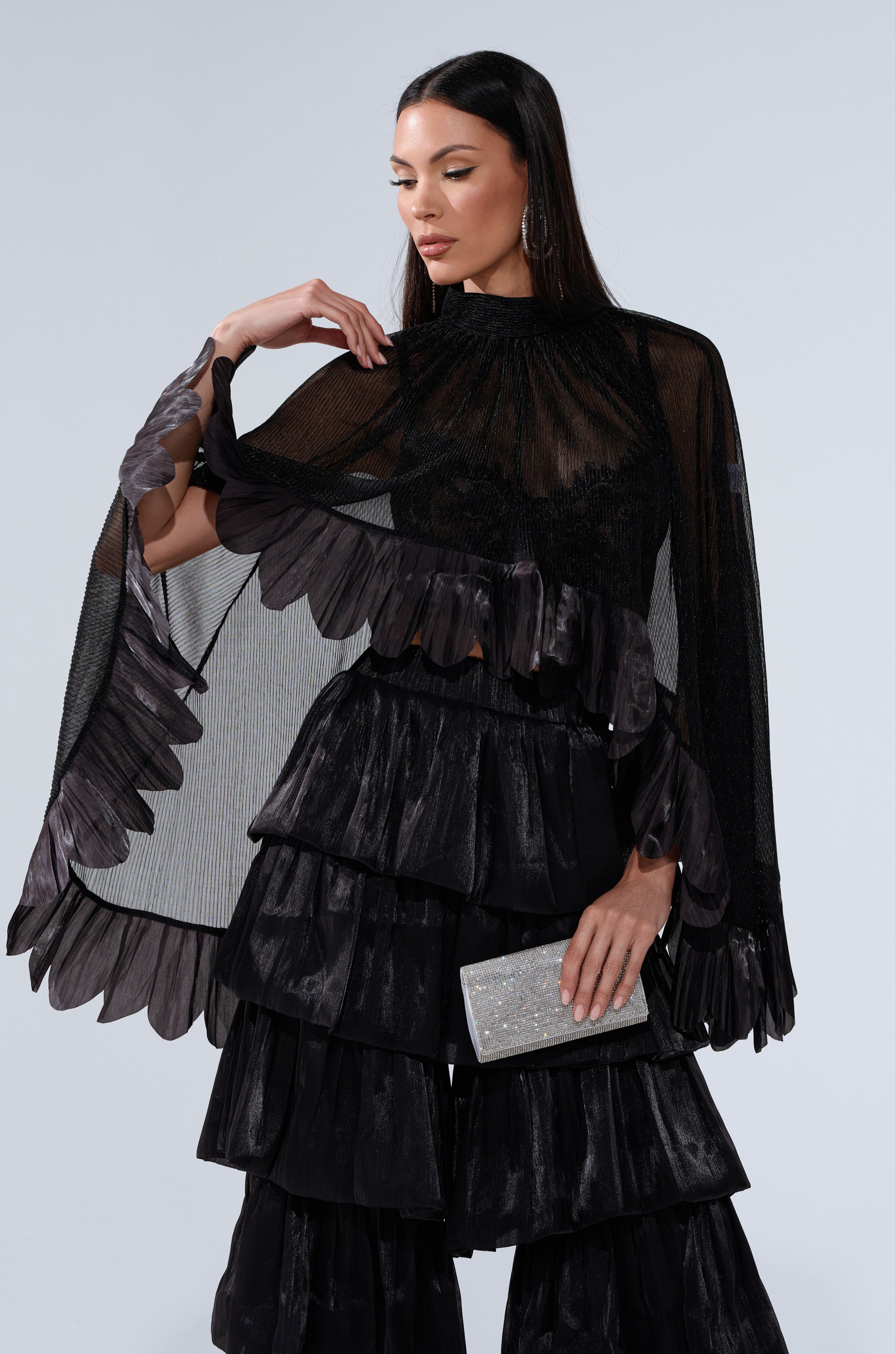 Wearing the BRIGHT LIFE BLOUSE with sheer plissé fabric and petal-like edges, paired with layered ruffled black pants, she holds a silver glitter clutch and poses gracefully against a plain light gray background.