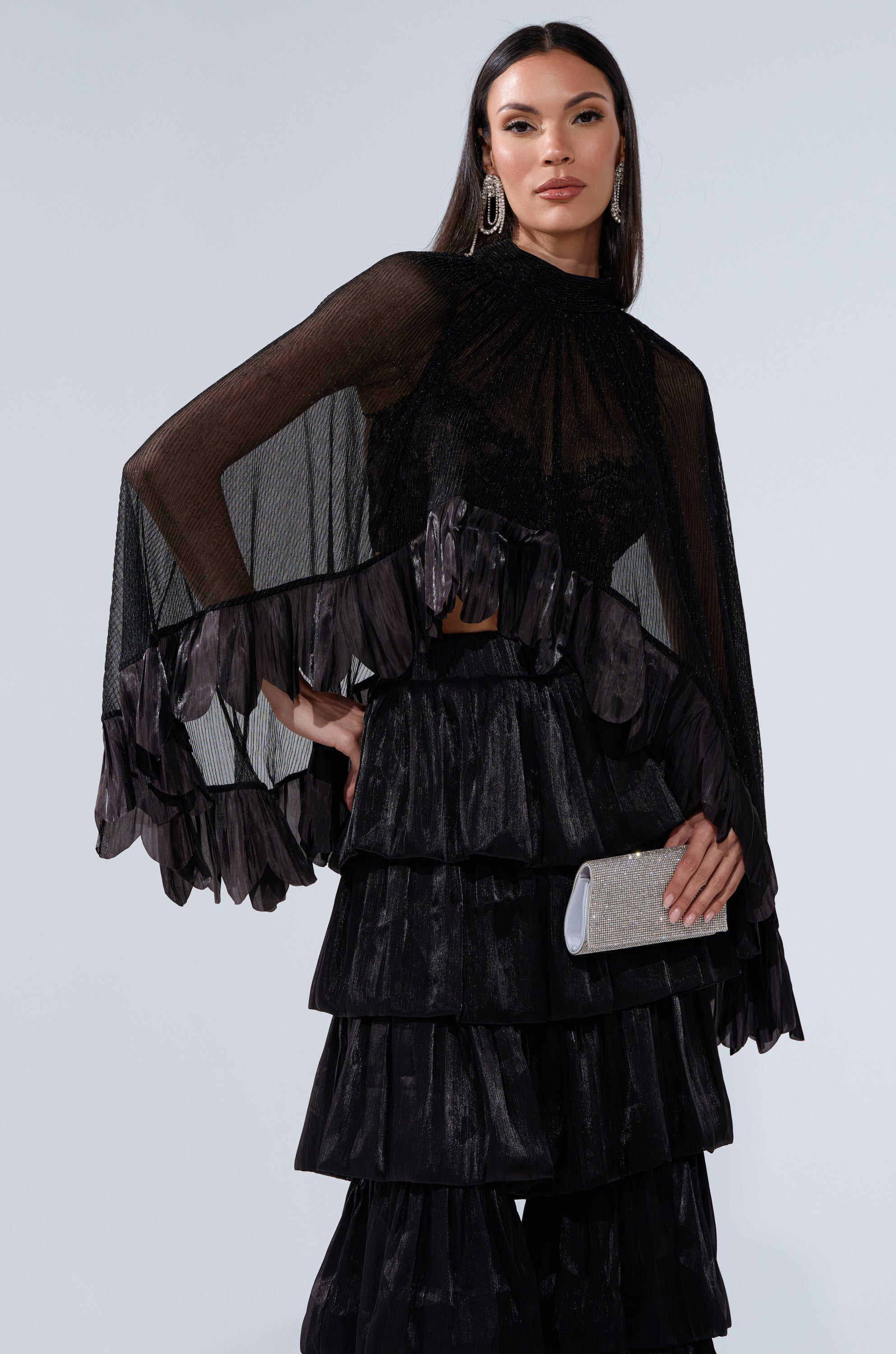 A woman with long dark hair and statement earrings poses against a plain light background, wearing the BRIGHT LIFE BLOUSE—a sheer plissé top with ruffled edges—and tiered black velvet pants, holding a sparkly silver clutch.