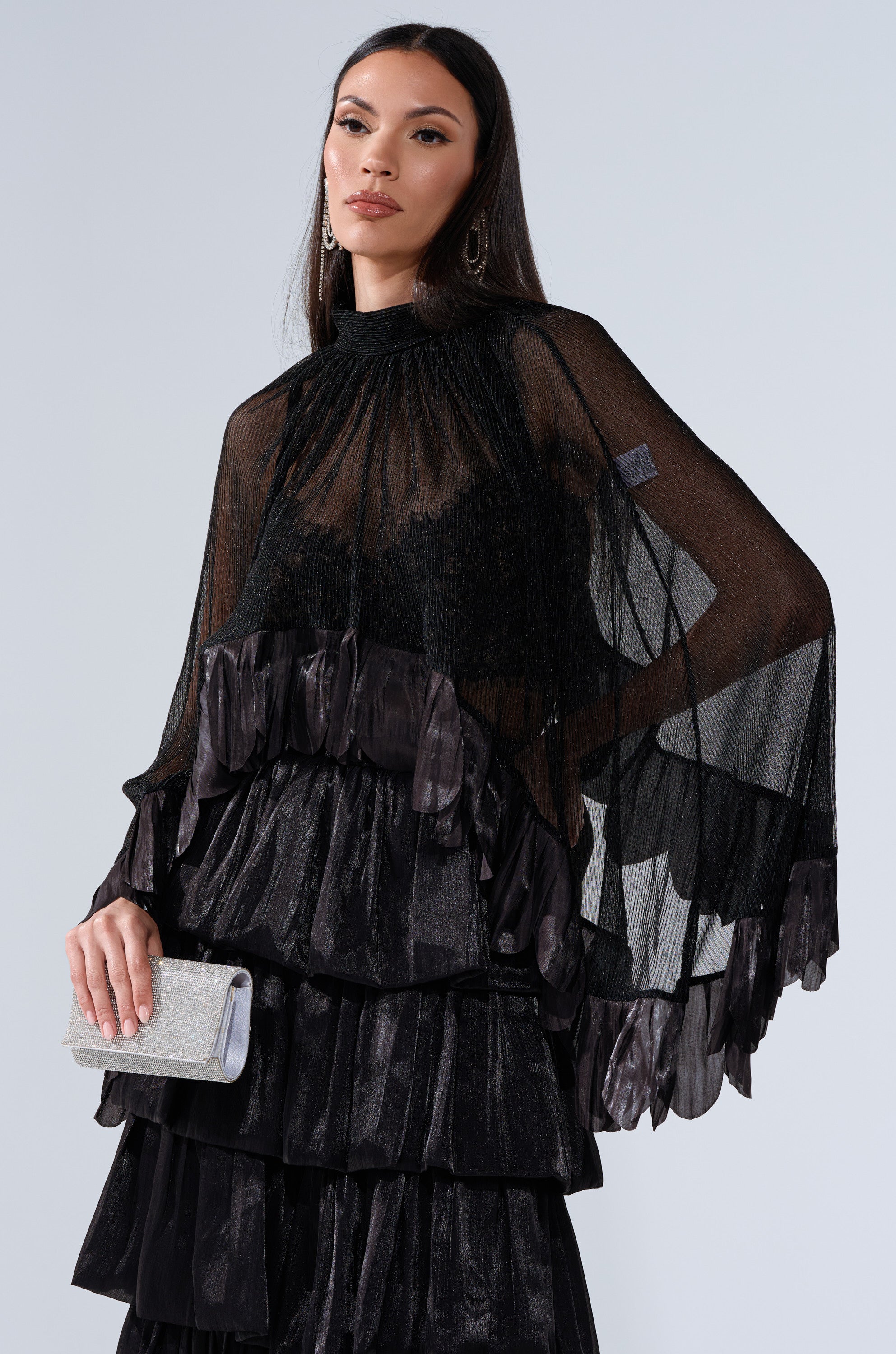 A woman wears the BRIGHT LIFE BLOUSE—a sheer plisse top with ruffled, tiered details—paired with a sparkly silver clutch. Her long straight hair and drop earrings stand out against a plain background.