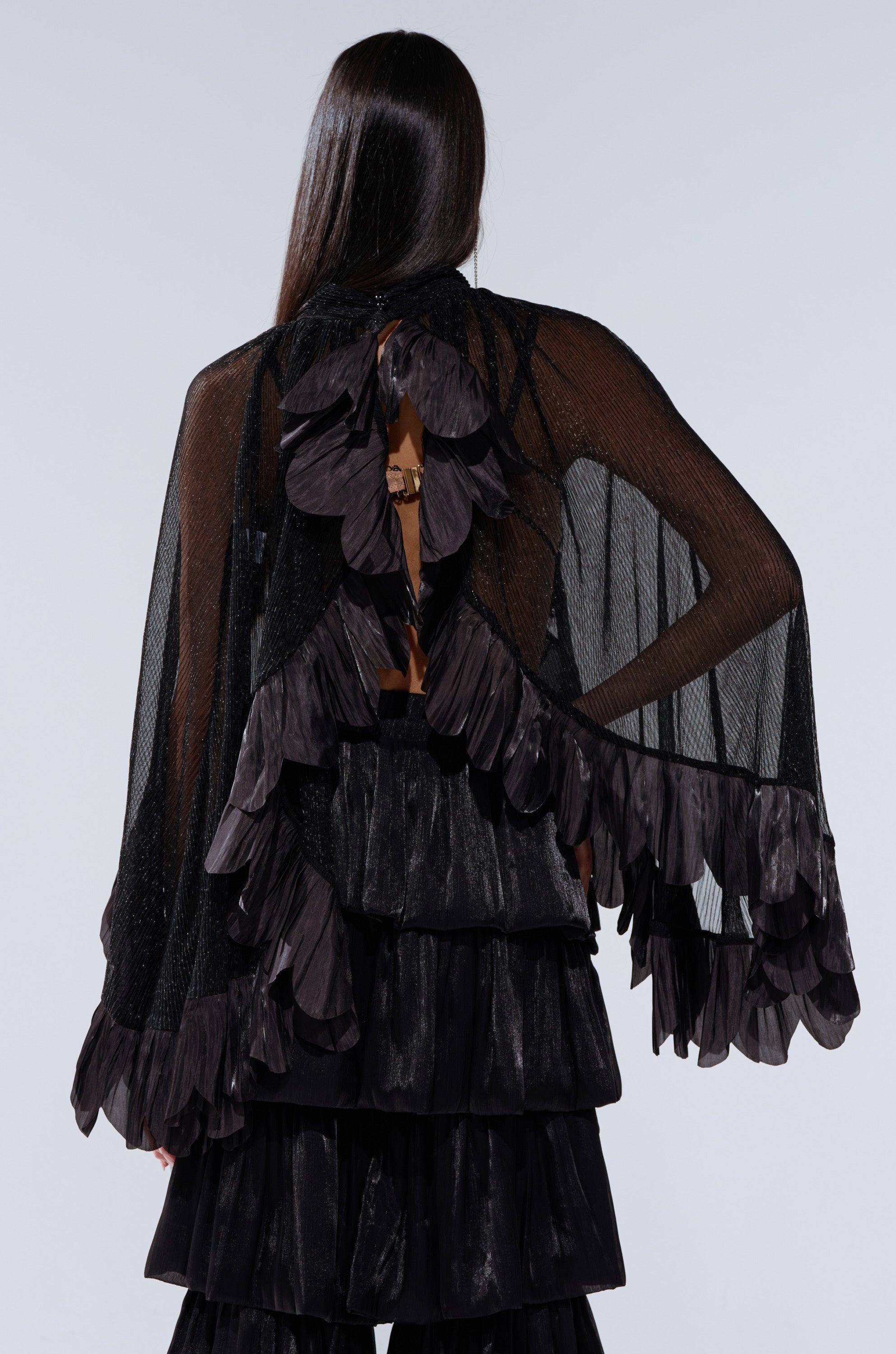 A person with long dark hair wears the BRIGHT LIFE BLOUSE, a sheer plissé top with layered ruffles and leaf details on the back, paired with matching tiered ruffle pants. Photo is taken from behind against a plain background.