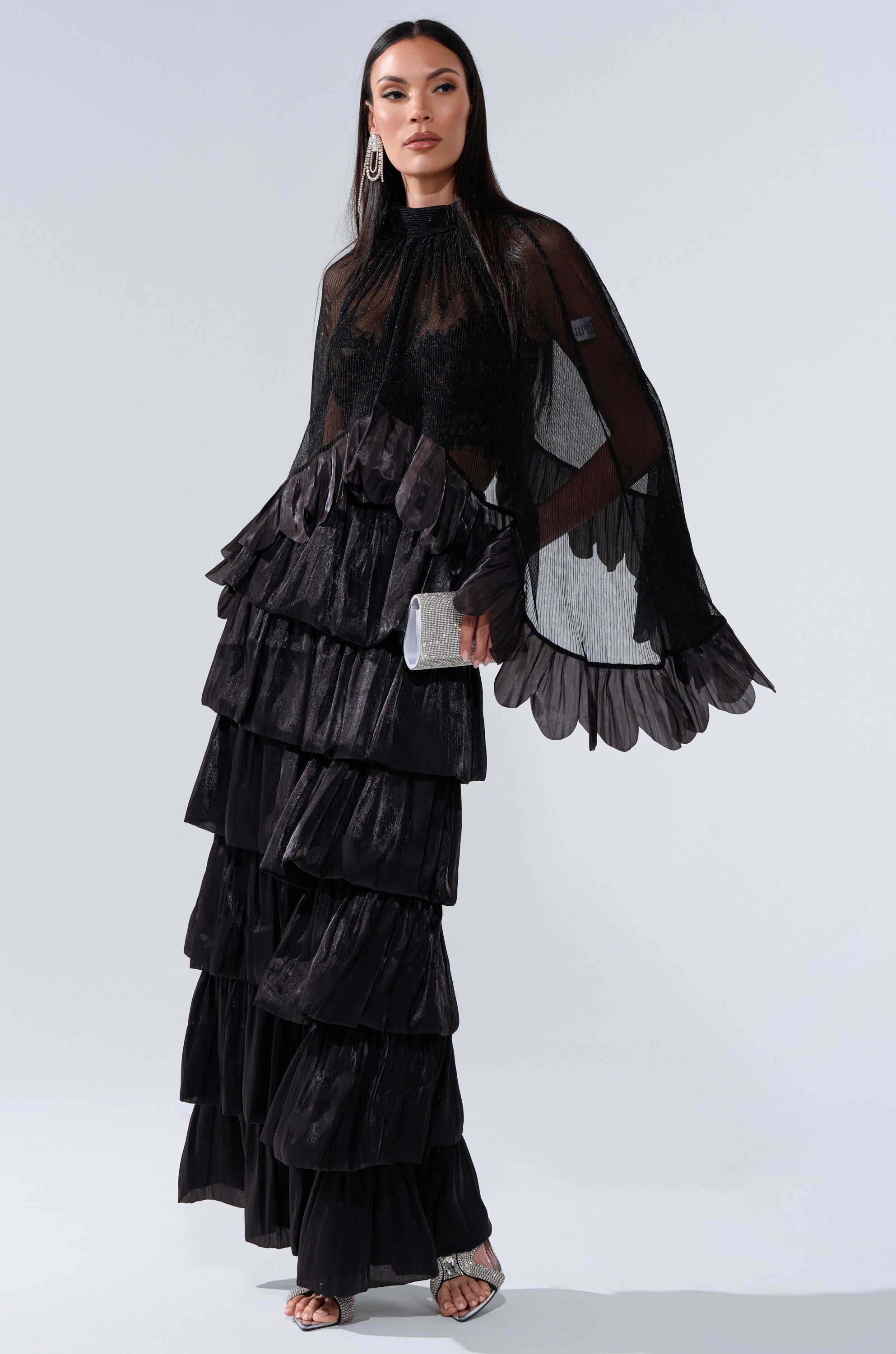 A woman poses in the BRIGHT LIFE BLOUSE, a sheer plissé top with scalloped edges, paired with a tiered ruffled black skirt. She accessorizes with a silver clutch and silver sandals against a plain light background.