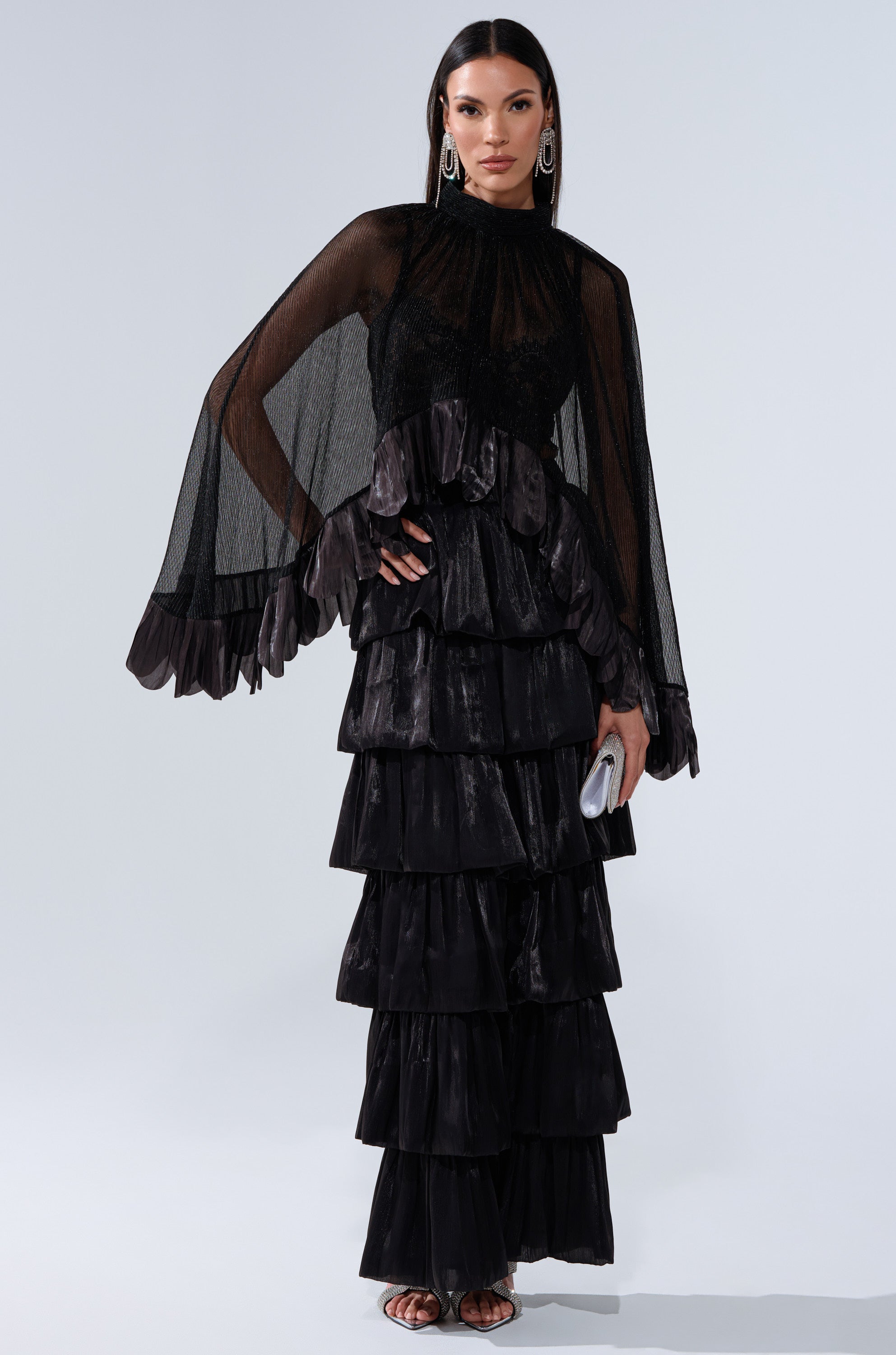 Wearing the BRIGHT LIFE BLOUSE with sheer plissé fabric and dramatic bell sleeves, she pairs it with a tiered black ruffle skirt, a silver clutch, large hoop earrings, and straight dark hair.