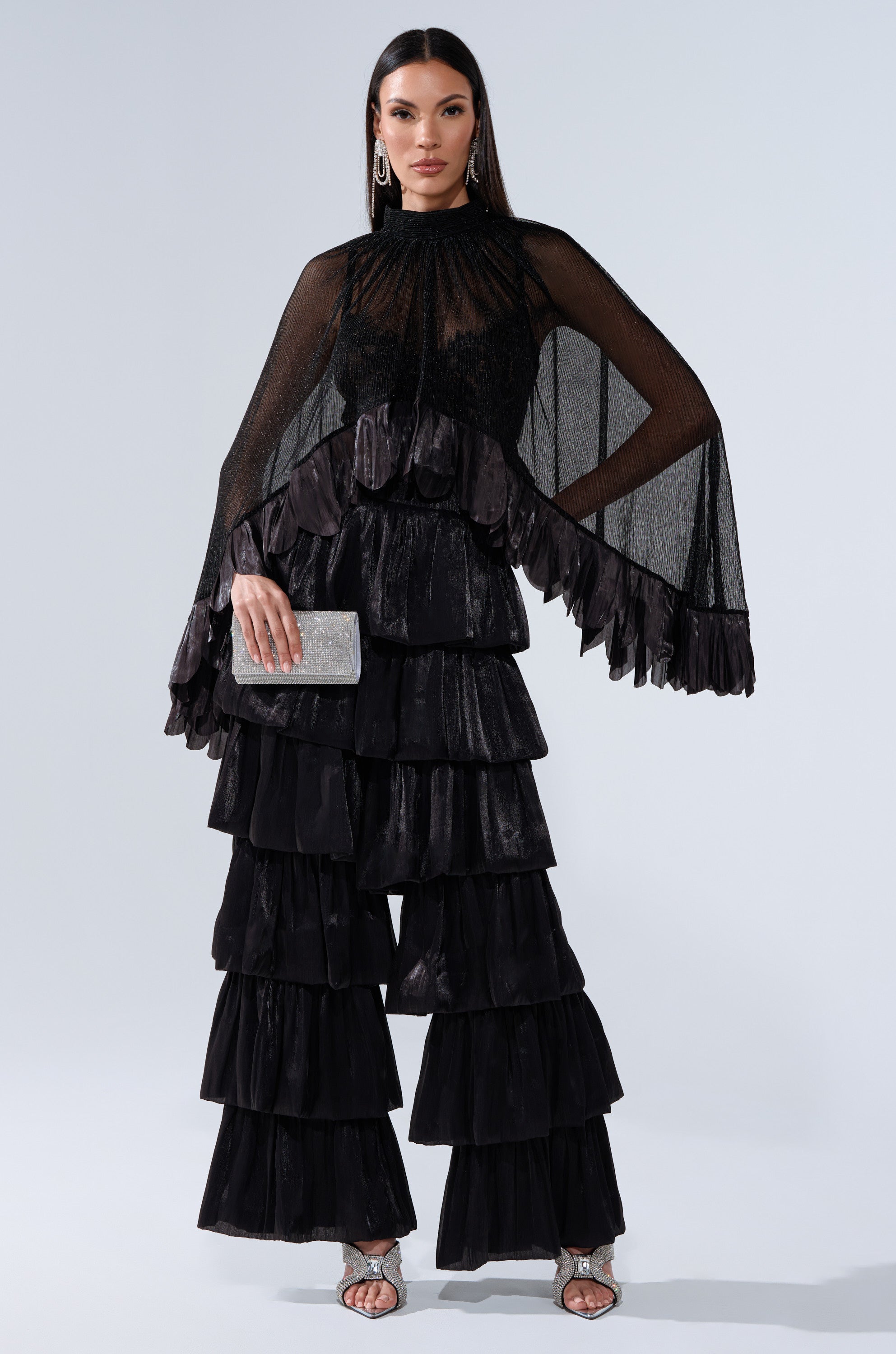 A woman in the dramatic black sheer BRIGHT LIFE BLOUSE with layered ruffle pants and cape-style top holds a sparkly silver clutch. She wears silver heels and has long, straight hair.