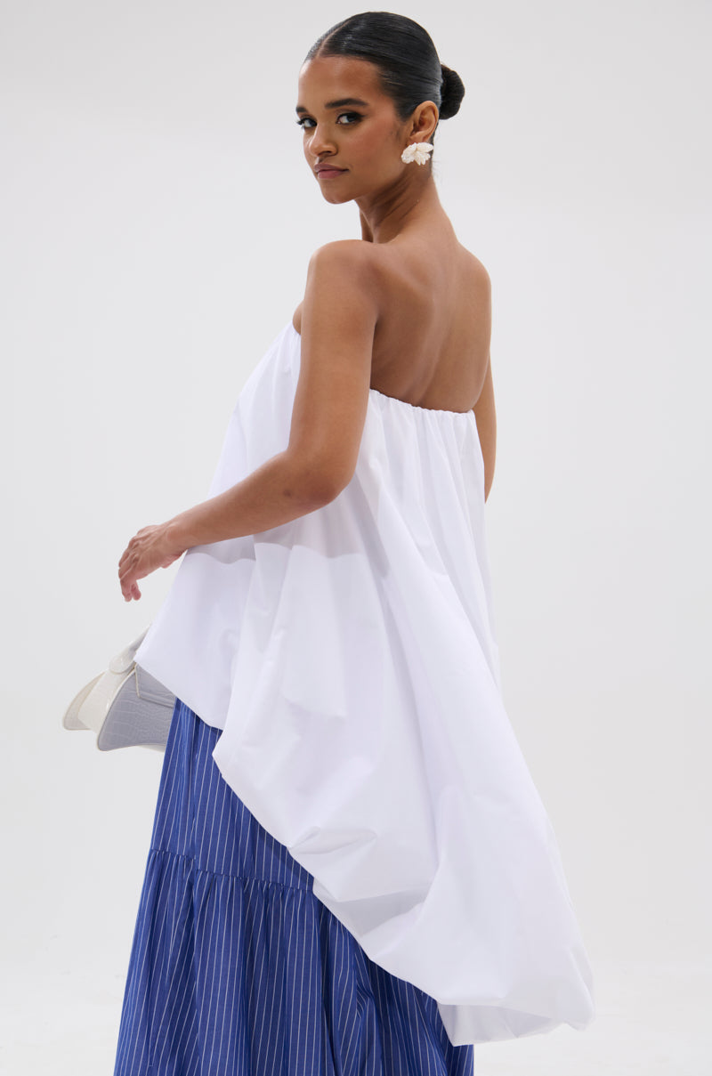 A woman poses in the OFF THE SHOULDER HI-LOW TOP with a long blue pleated skirt, holding a white handbag and wearing statement earrings against a plain light background, capturing modern minimalism.