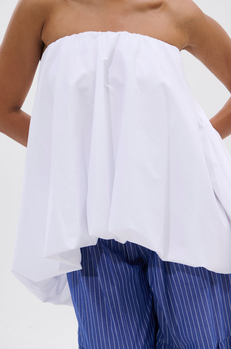 Wearing the OFF THE SHOULDER HI-LOW TOP and blue pinstriped pants, a person stands against a plain white background with bent arms and hands on hips.