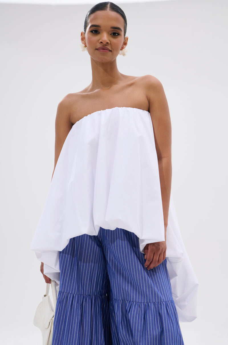 A woman embraces modern minimalism in the OFF THE SHOULDER HI-LOW TOP, paired with blue wide-legged pinstriped pants and a white bag, standing confidently against a plain light background.