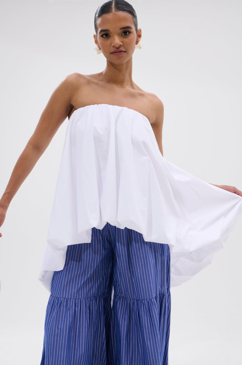 A woman models the OFF THE SHOULDER HI-LOW TOP with wide-leg blue pants featuring white stripes, standing against a plain background and holding out one side of her top for a modern minimalist look.