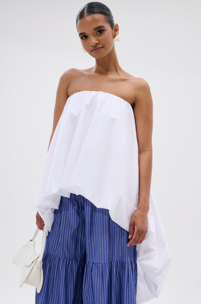 A woman in an OFF THE SHOULDER HI-LOW TOP and blue pinstriped wide-leg pants stands against a light background, holding a small white handbag and smiling gently, expressing modern minimalism.