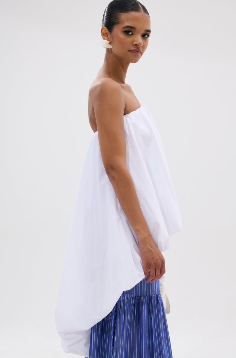 A woman in a modern minimal look wears the OFF THE SHOULDER HI-LOW TOP with an asymmetric hem over a blue pleated skirt, styled with pearl earrings and slicked-back hair against a neutral background.