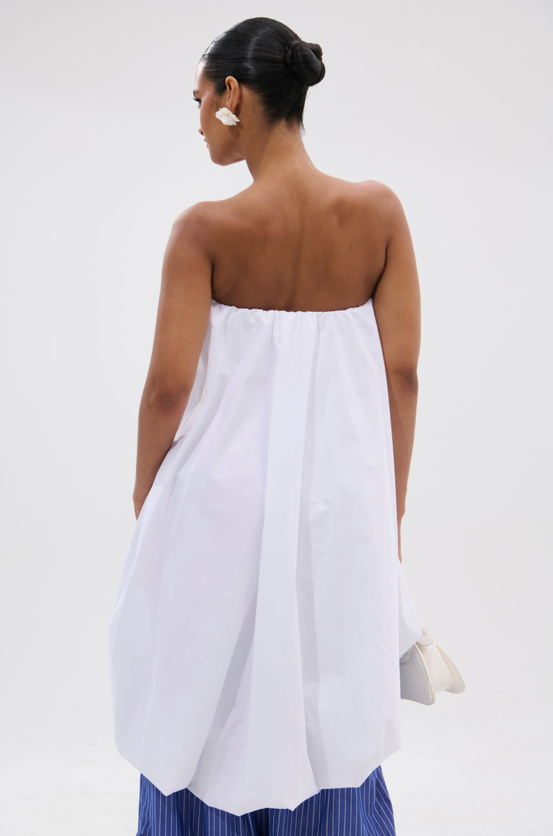 A woman with her hair in a bun wears an OFF THE SHOULDER HI-LOW TOP, featuring strapless white fabric, paired with blue pinstriped pants and white earrings. She stands facing away against a plain, minimalist light background.