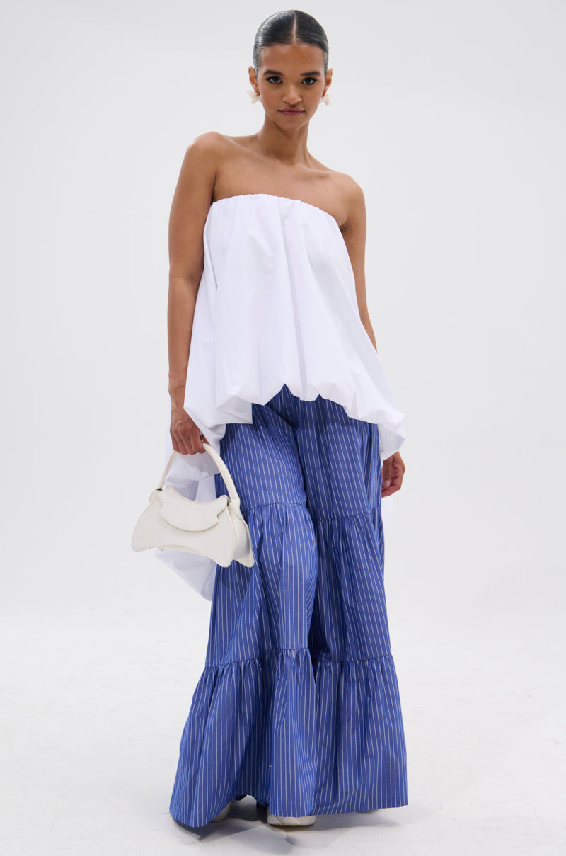 A woman poses in a minimal look, wearing an OFF THE SHOULDER HI-LOW TOP and blue, wide-leg tiered pinstripe pants. She holds a small white handbag and has her hair back against a plain white background.