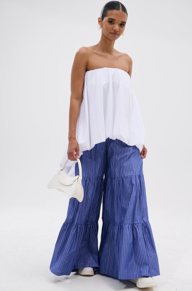 A woman stands against a white background wearing the OFF THE SHOULDER HI-LOW TOP and wide-legged, tiered blue striped pants. She holds a small white handbag, wears white shoes, and has her hair tied back.