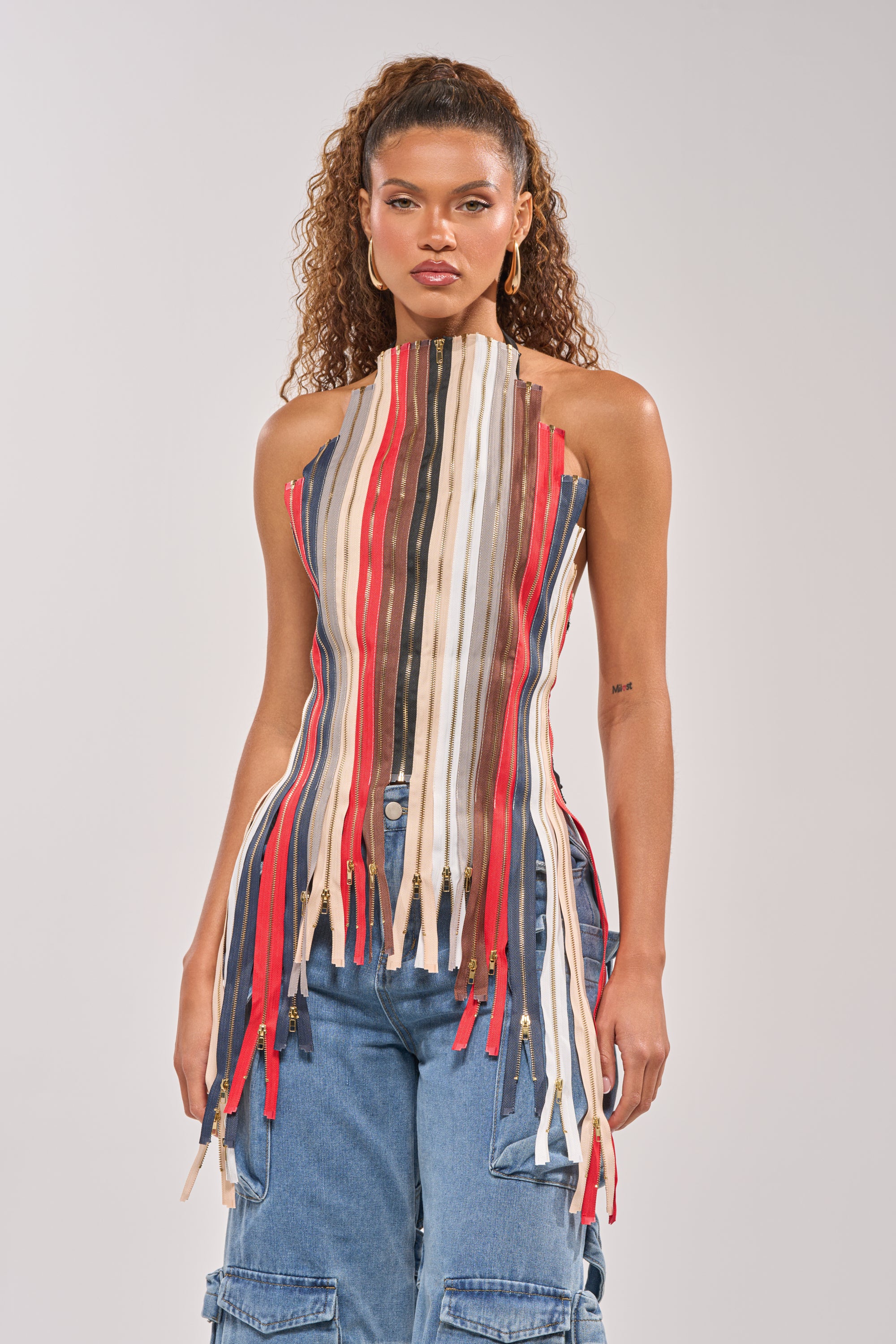 A woman with curly hair models the ZIPPED UP HALTER TOP, a corset-style piece with red, beige, white, and brown vertical strips and zipper pulls, paired with blue cargo jeans—making for standout festival wear against a plain light background. 0