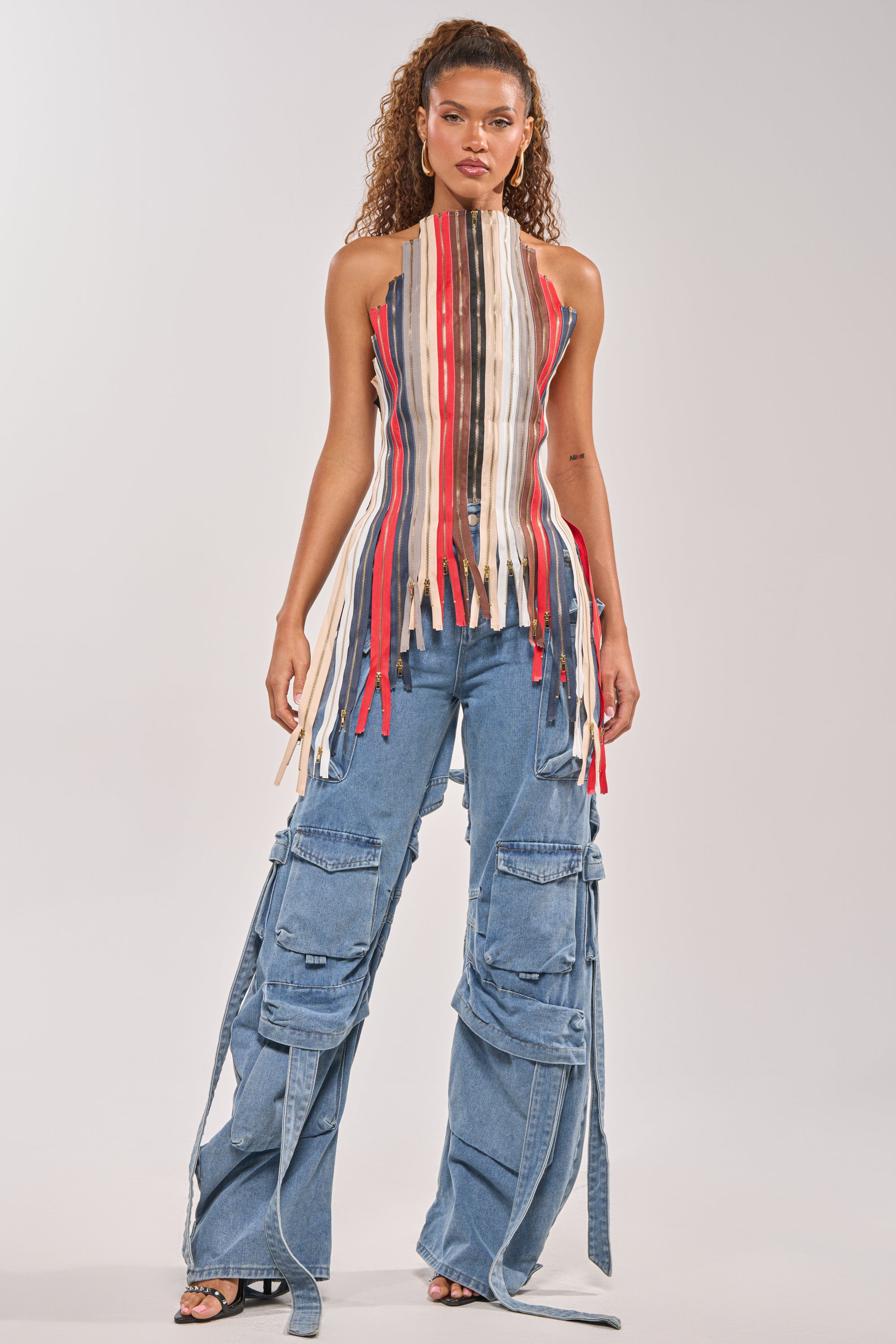 A woman with curly hair and a neutral look wears the ZIPPED UP HALTER TOP, featuring long vertical red, white, and tan strips, paired with loose blue cargo jeans with multiple pockets and straps. 9