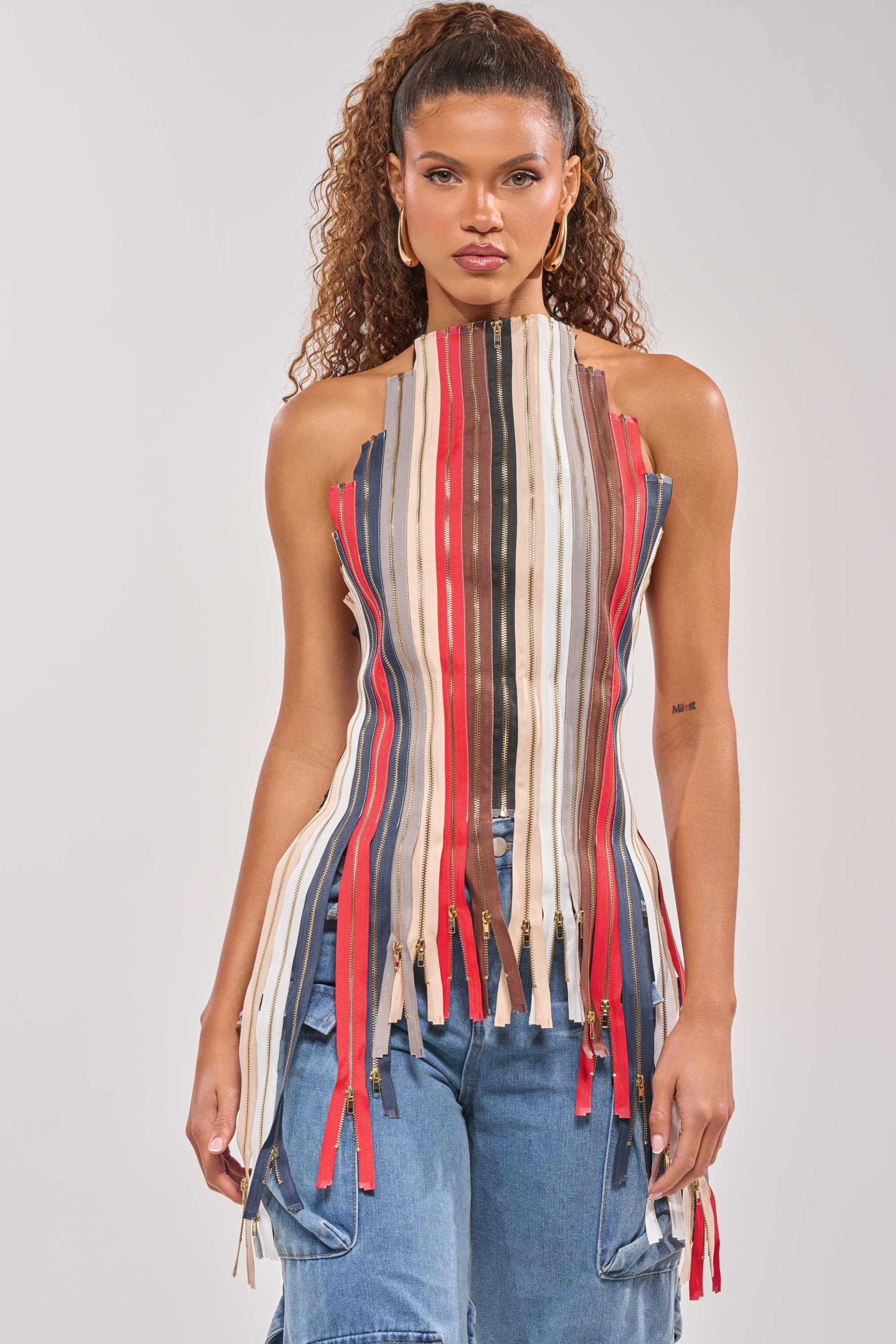 A woman with curly hair models the ZIPPED UP HALTER TOP, a sleeveless, high-neck style with vertical multicolored strips and hanging ends—ideal for festivals—paired with blue jeans, standing against a plain backdrop. 1