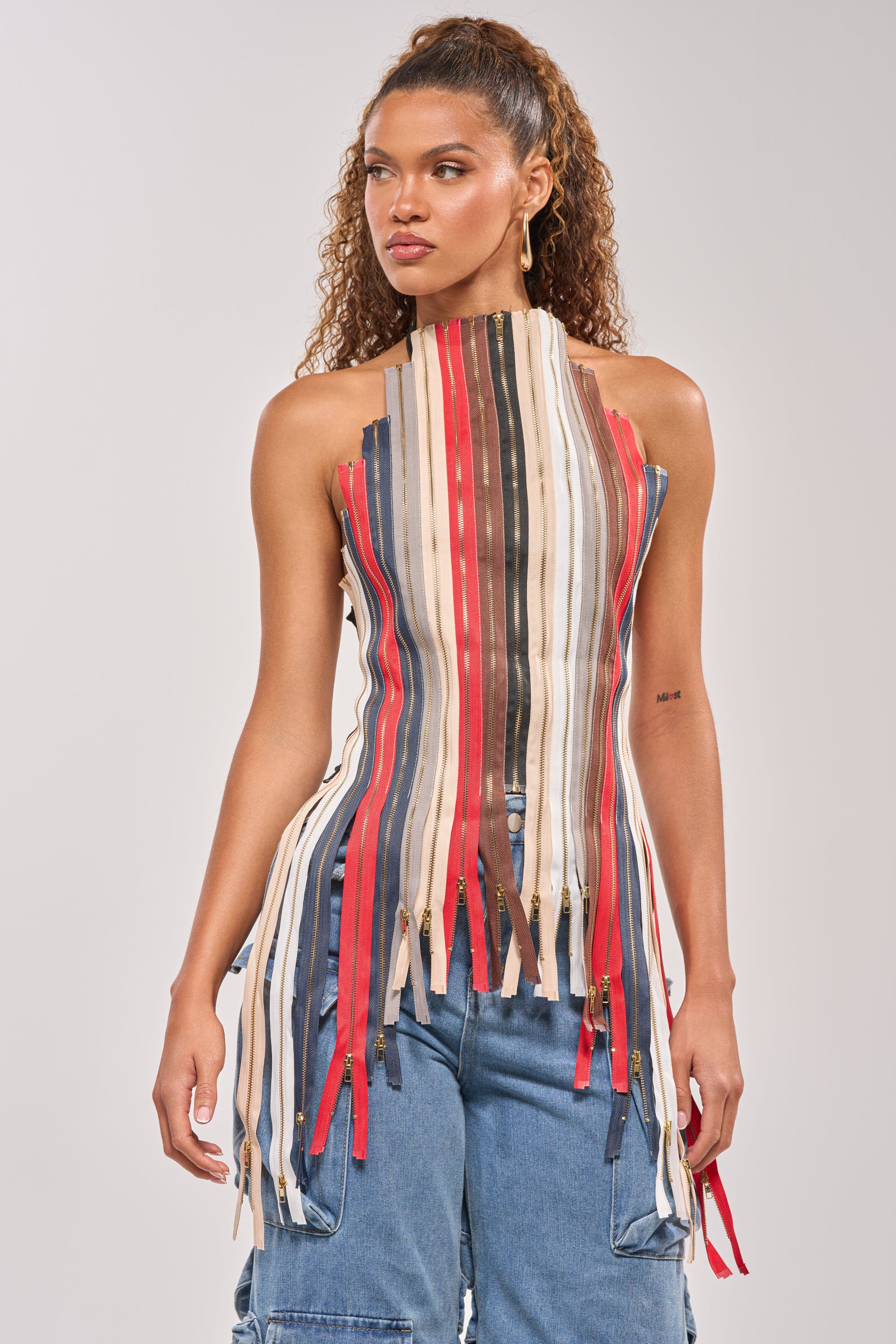 A woman wears the ZIPPED UP HALTER TOP, featuring vertical, multicolored fabric strips and a zipper front, paired with blue cargo jeans. Her curly hair is styled in a high half-ponytail with gold hoop earrings—ideal for festival wear. 2