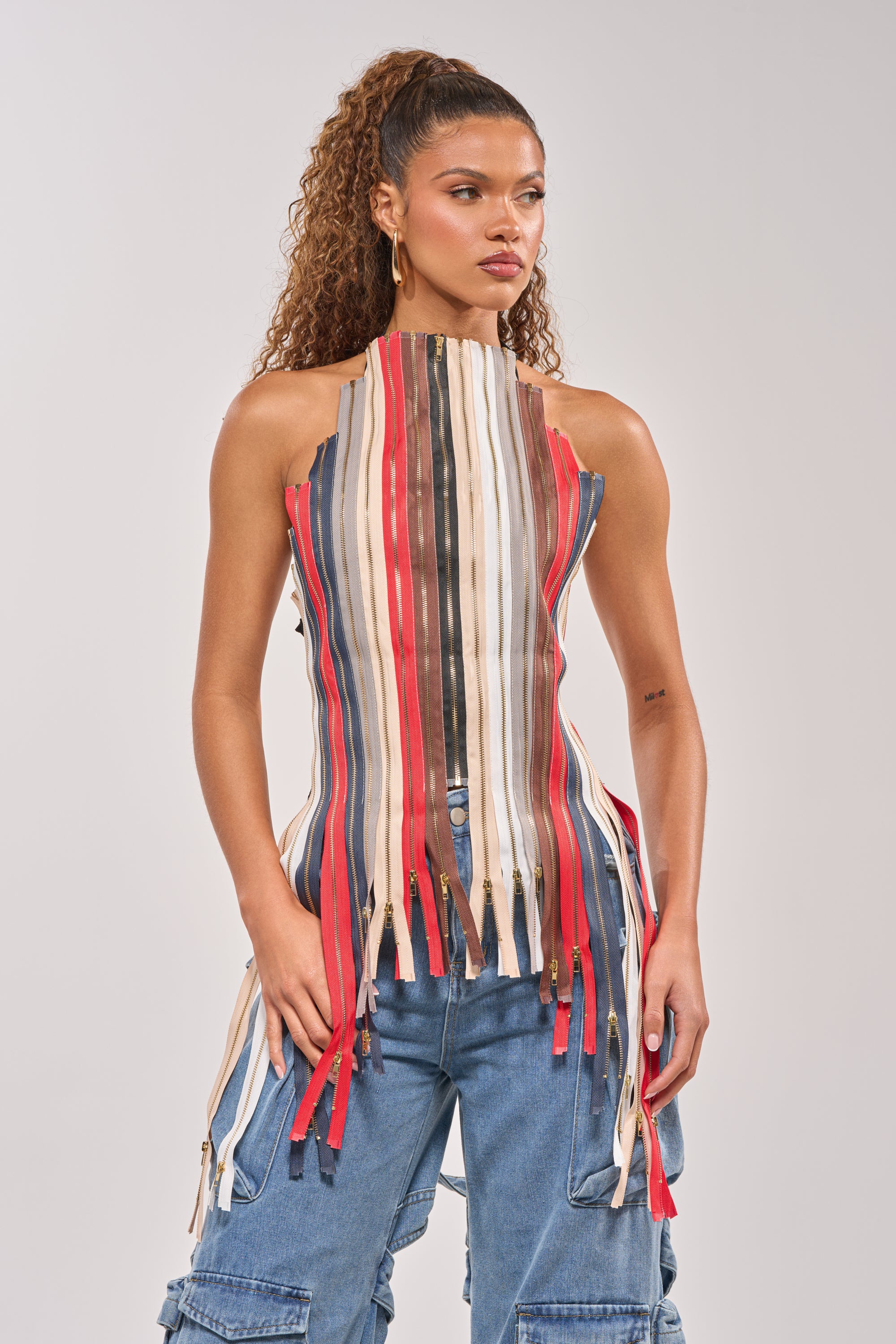 A woman with curly hair in a high ponytail wears the ZIPPED UP HALTER TOP, a sleeveless, high-neck piece with vertical multicolored stripes and fringe hem, paired with blue cargo pants. She stands confidently against a plain background. 3