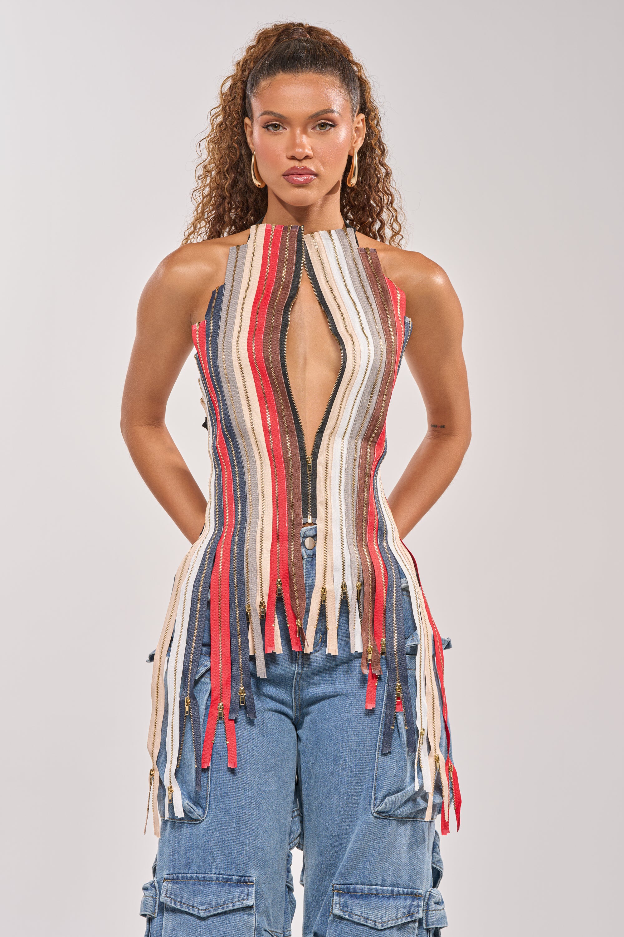 A woman with curly hair wears the ZIPPED UP HALTER TOP, featuring vertical multicolored stripes, a zipper front, and deep V neckline, paired with blue cargo jeans. She stands against a plain light background. 4