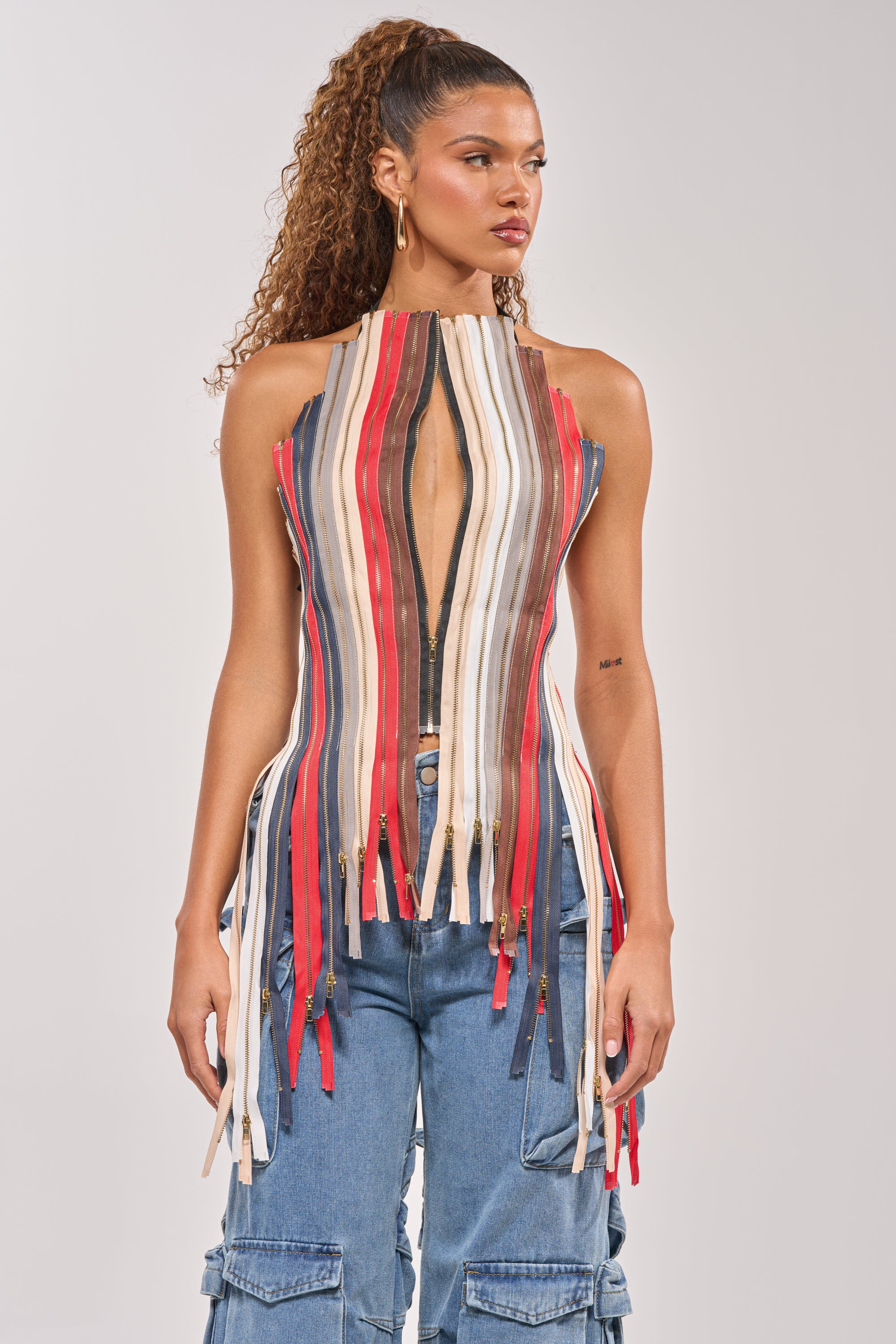 A woman wears the ZIPPED UP HALTER TOP, a festival-ready corset with a halter neckline, vertical multicolored stripes, and zippers. She pairs it with blue cargo jeans and sports a high curly ponytail against a plain background. 5
