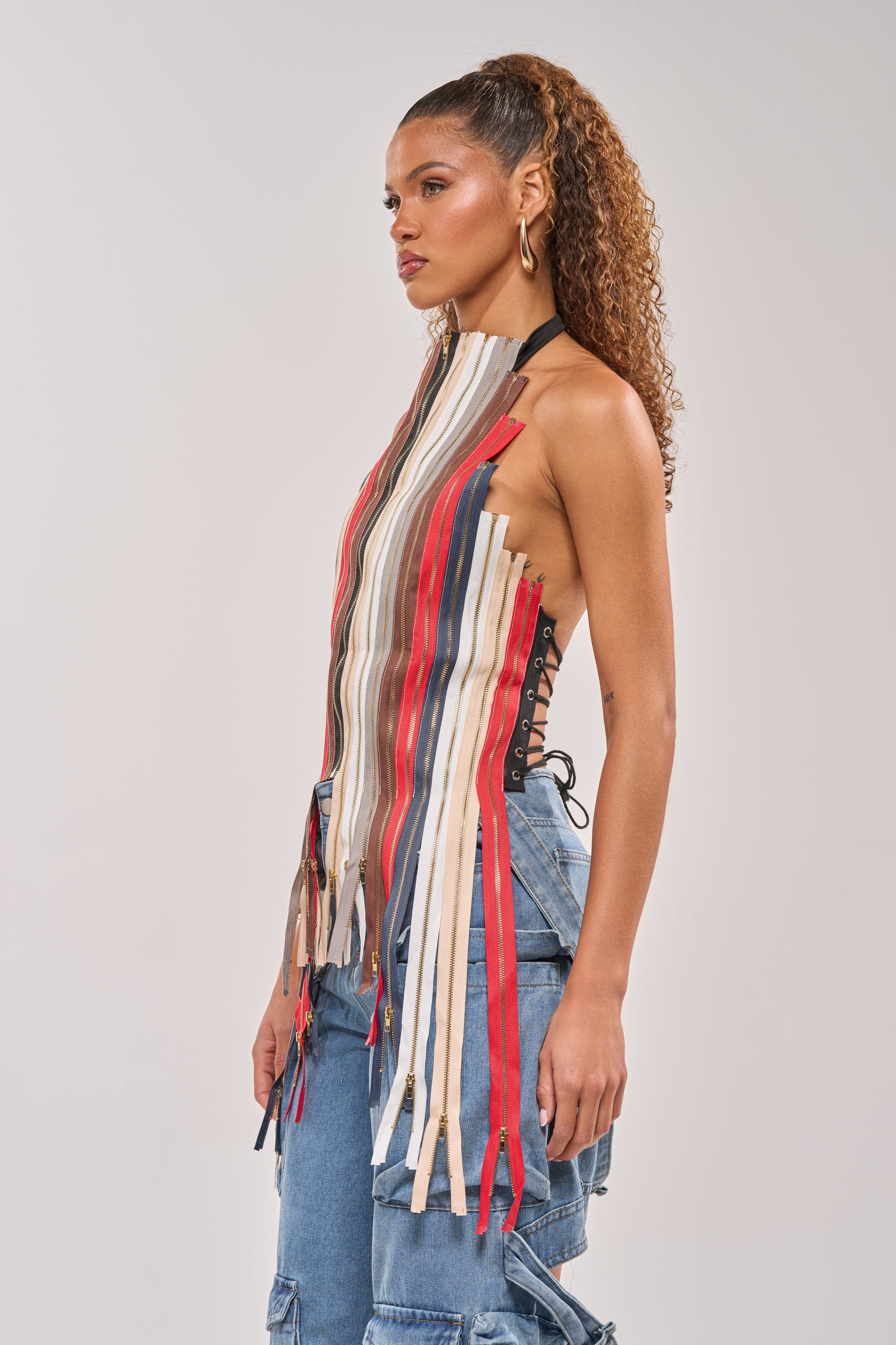 A woman with curly hair in a high ponytail wears gold hoop earrings and the ZIPPED UP HALTER TOP—a sleeveless, multicolored vertical-striped corset—paired with blue lace-up side denim pants, perfect for festivals. She stands in profile against a plain background. 6