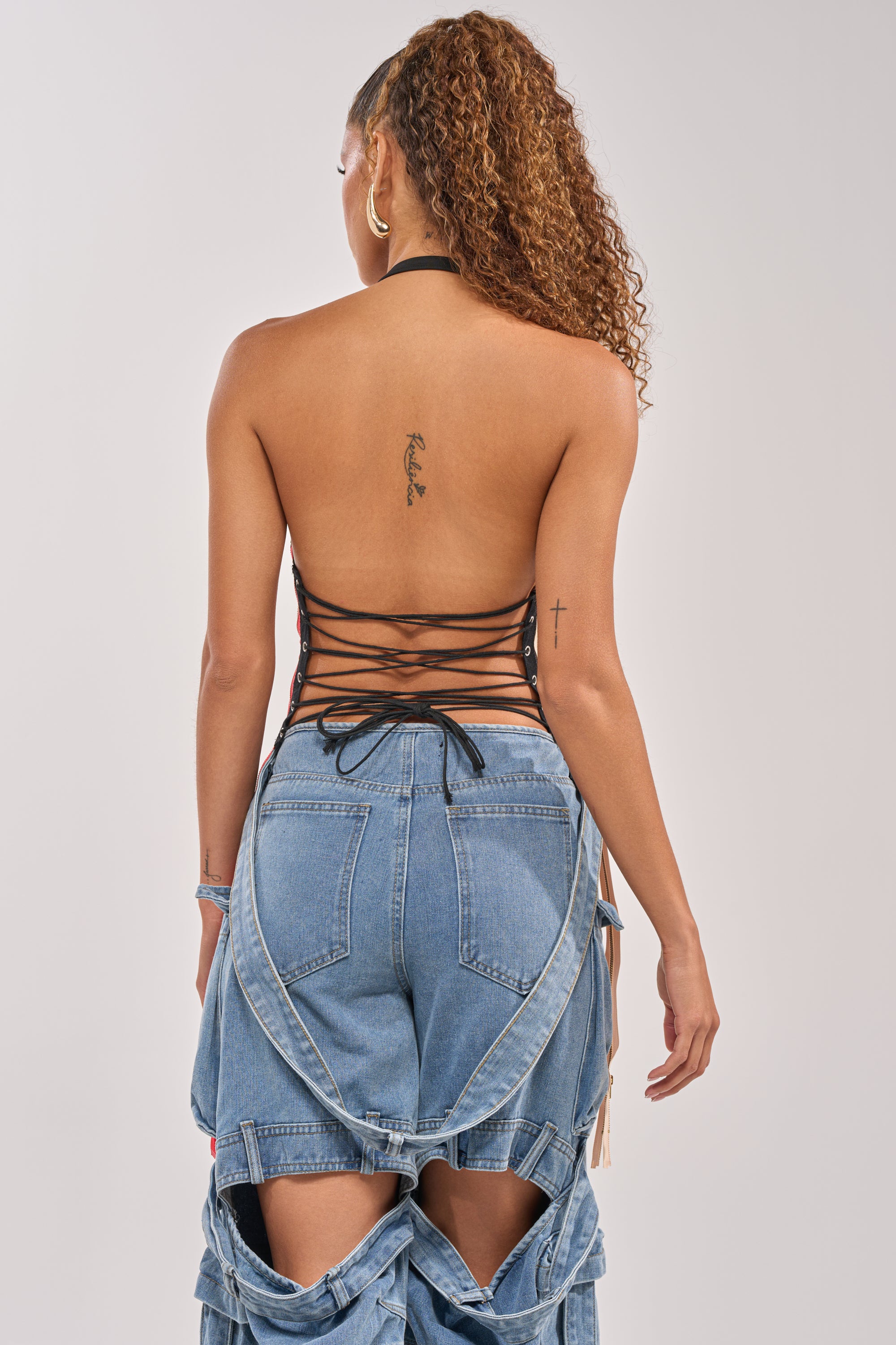 A woman with curly hair in a high ponytail wears the ZIPPED UP HALTER TOP and baggy blue jeans with loose suspenders—festival-ready style. Facing away, she reveals a small tattoo on her upper back. 7