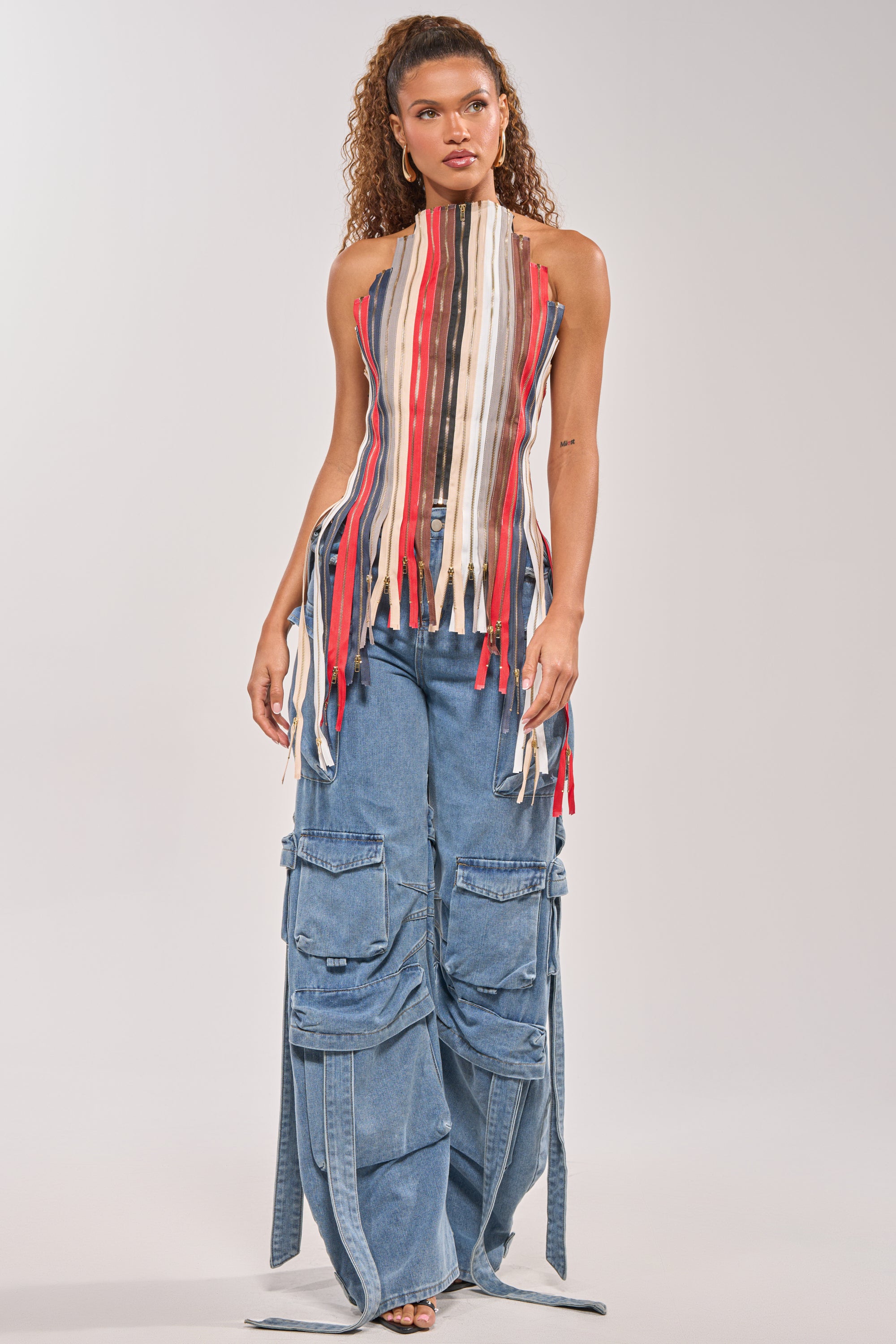 A woman wears the ZIPPED UP HALTER TOP, a festival-ready, sleeveless piece with vertical red, white, black, and beige strips, paired with high-waisted blue wide-leg cargo jeans with large pockets and extra denim straps. 8