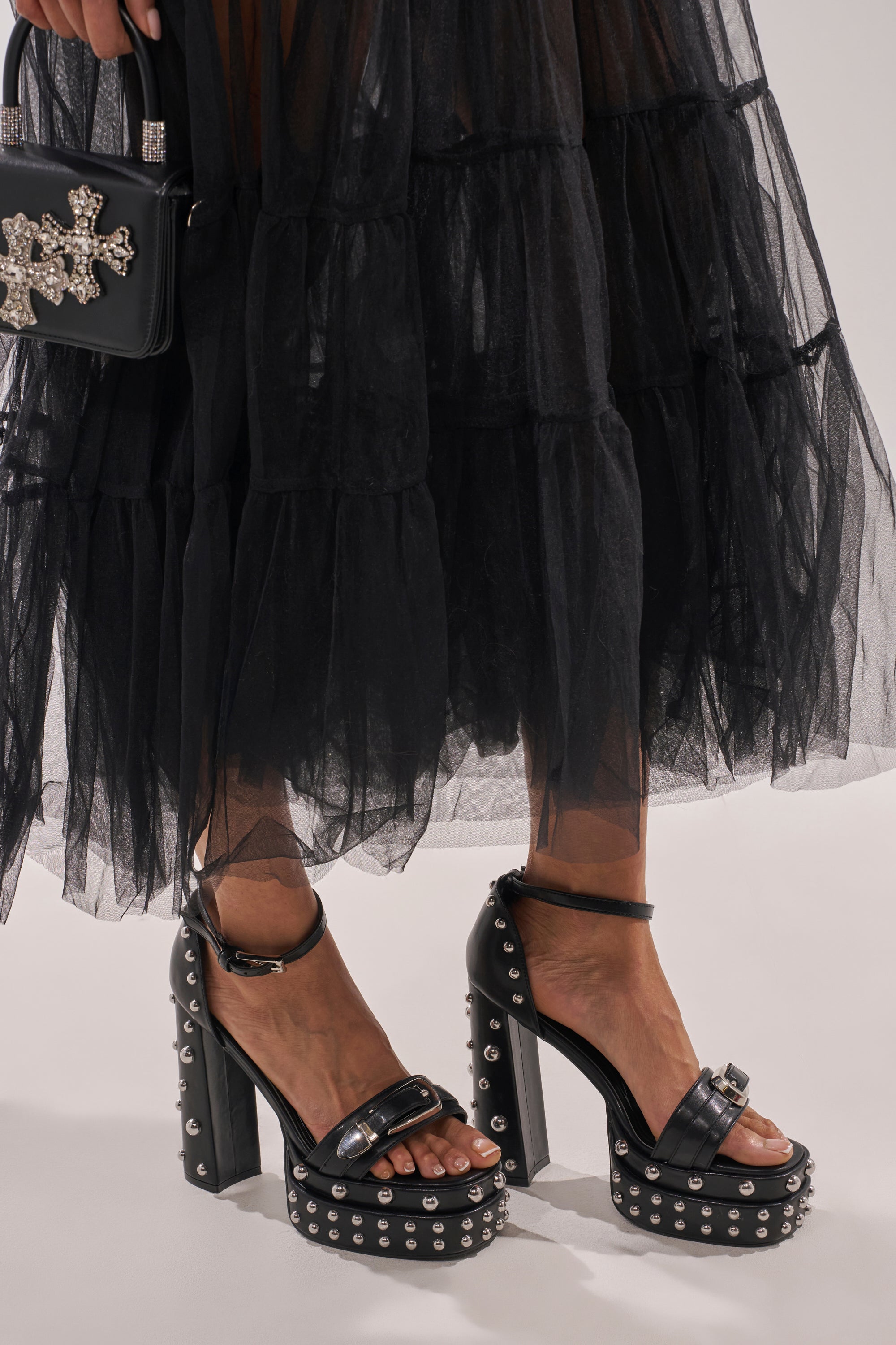 Wearing a sheer black tulle dress, she holds a sparkling embellished black handbag and wears AZALEA WANG EUGENIJA BLACK CHUNKY EMBELLISHED STUDDED SANDALS with a platform sole and block heel. 0