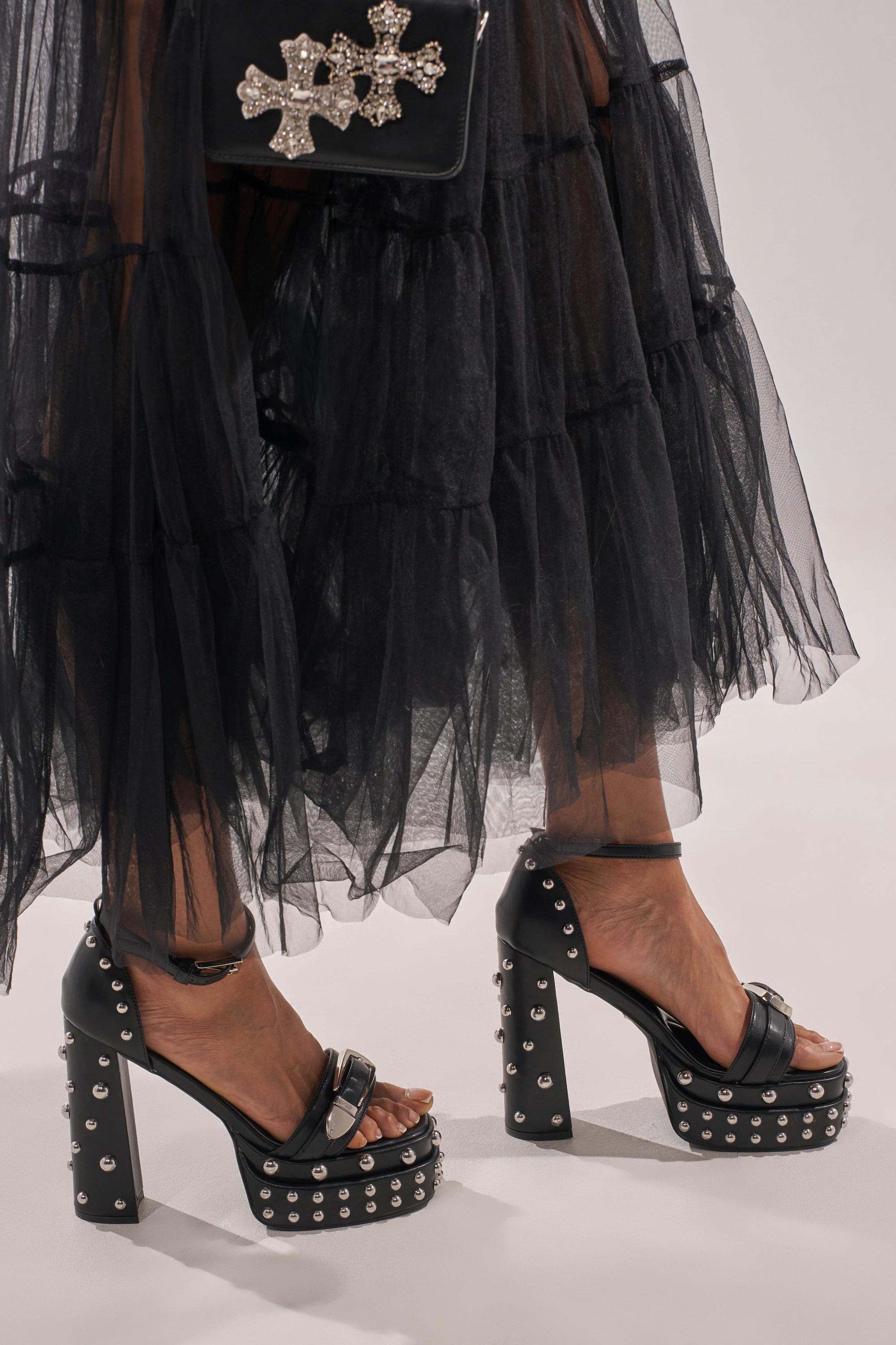 A person wears a sheer black ruffled skirt, holds a black purse with silver crosses, and completes the edgy look with the AZALEA WANG EUGENIJA BLACK CHUNKY EMBELLISHED STUDDED SANDAL. 1