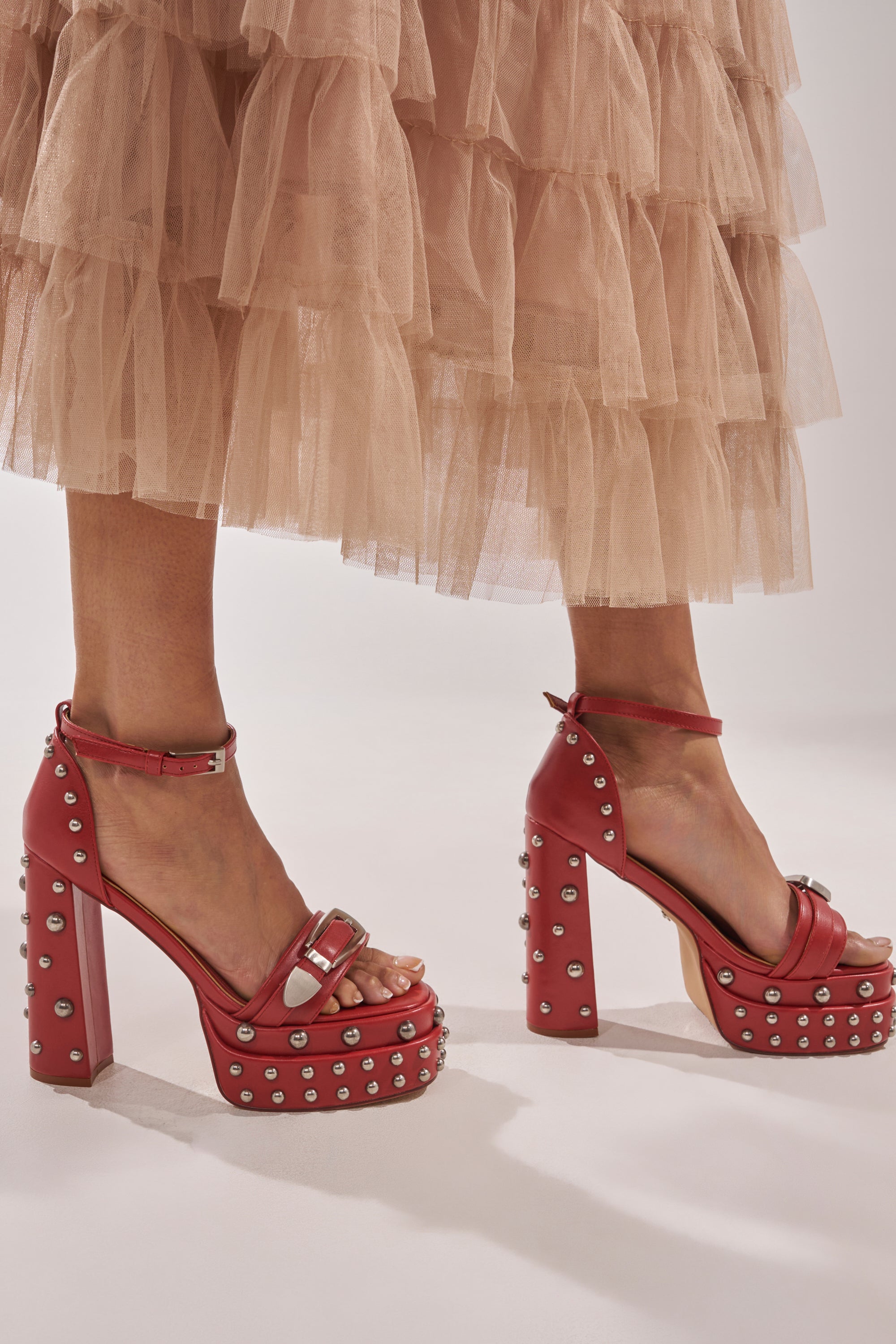 A person in a tiered beige tulle skirt and AZALEA WANG EUGENIJA RED CHUNKY EMBELLISHED STUDDED SANDALS with faux leather ankle straps stands on a light-colored surface. 0