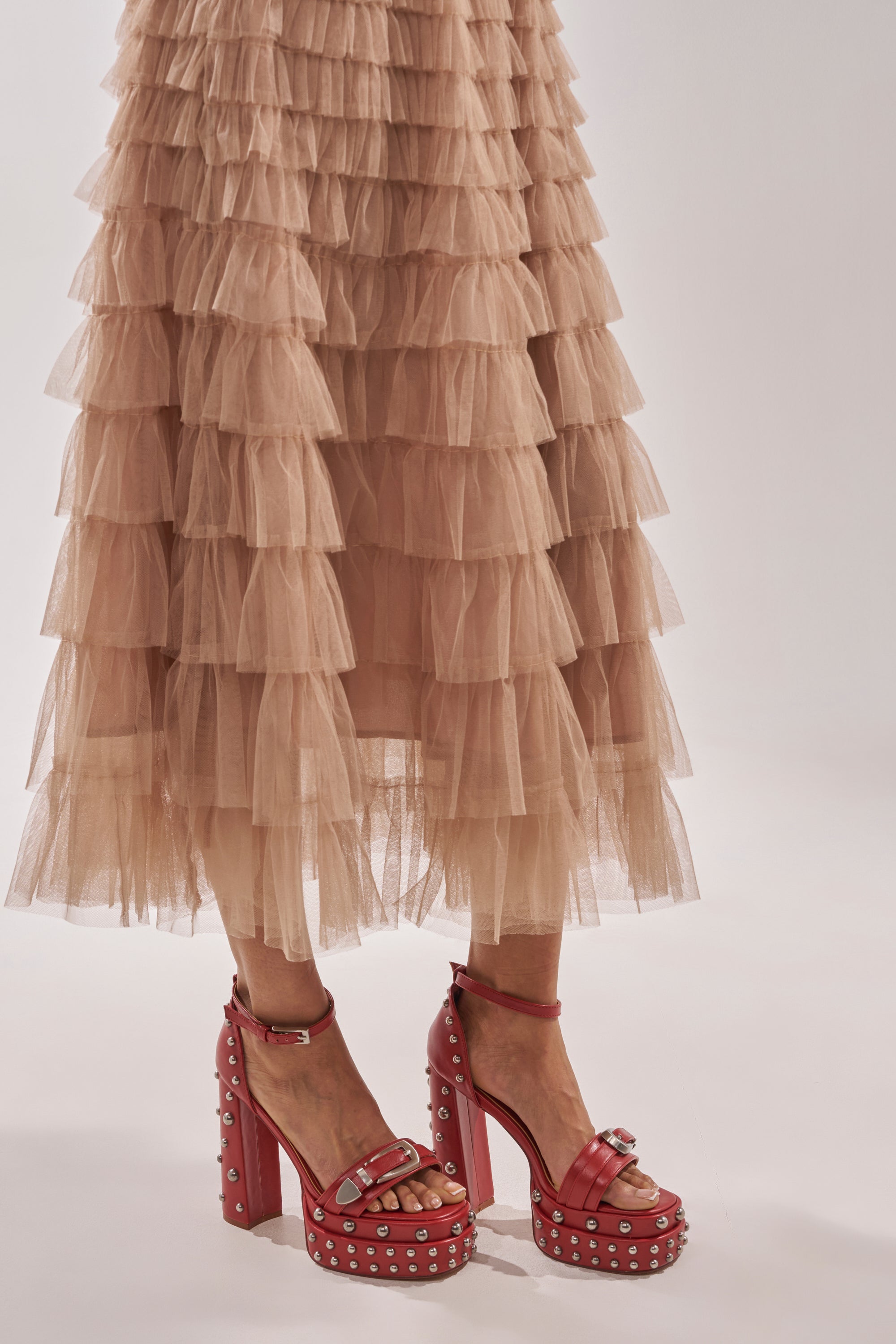 A person styles a layered beige tulle skirt with AZALEA WANG EUGENIJA Red Chunky Embellished Studded Sandals, featuring faux leather and platform heels, standing against a light background. 1