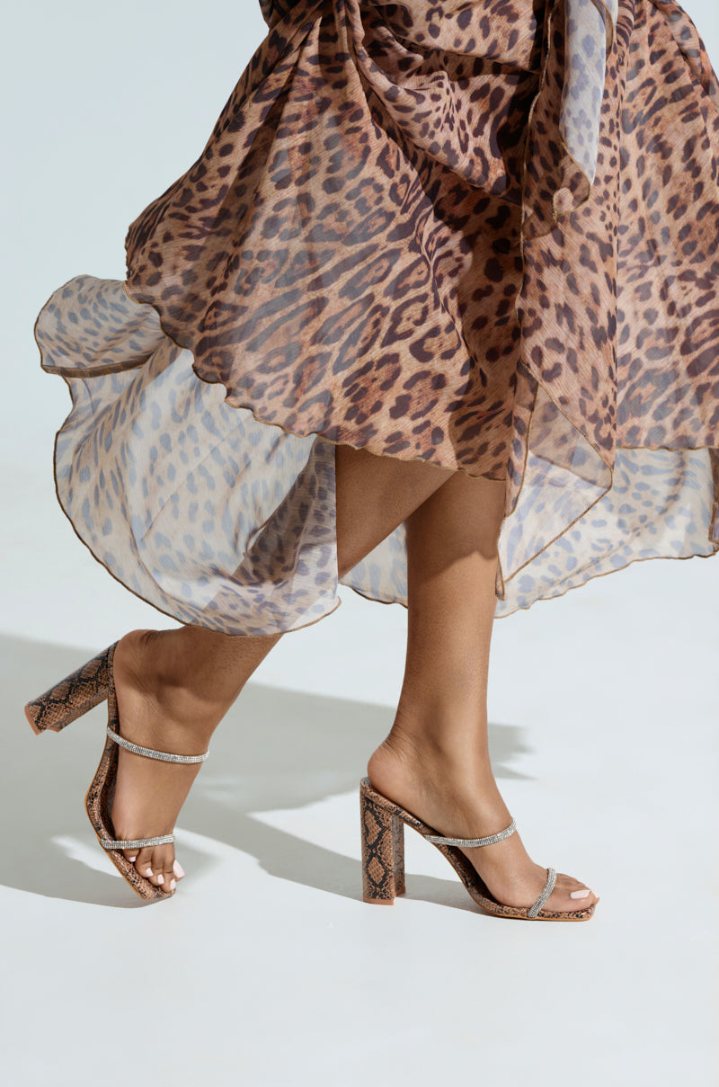 A person walks gracefully in a flowing leopard-print dress, wearing clear AZALEA WANG Eulalia Snake Slip On Sandals with rhinestone straps. The dress swirls around their legs against a light background, while the chunky block heel shimmers with each step.