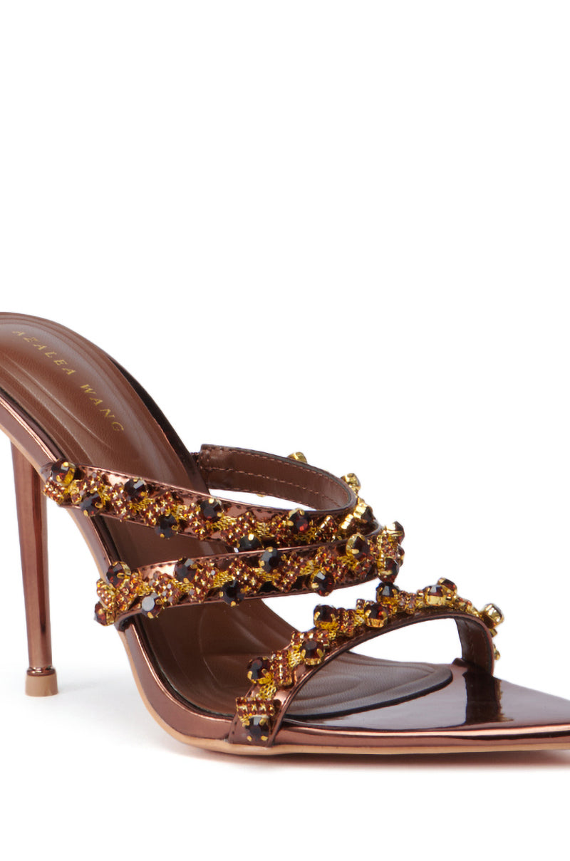 Introducing the AZALEA WANG EUREKA BRONZE EMBELLISHED STILETTO SANDAL, a luxurious brown high-heeled sandal featuring a faux leather upper adorned with gold and amber decorative jewels on the straps. This elegant shoe boasts a slender heel, while its footbed proudly displays the designer's name in gold letters, enhancing its sophisticated and glamorous appeal.