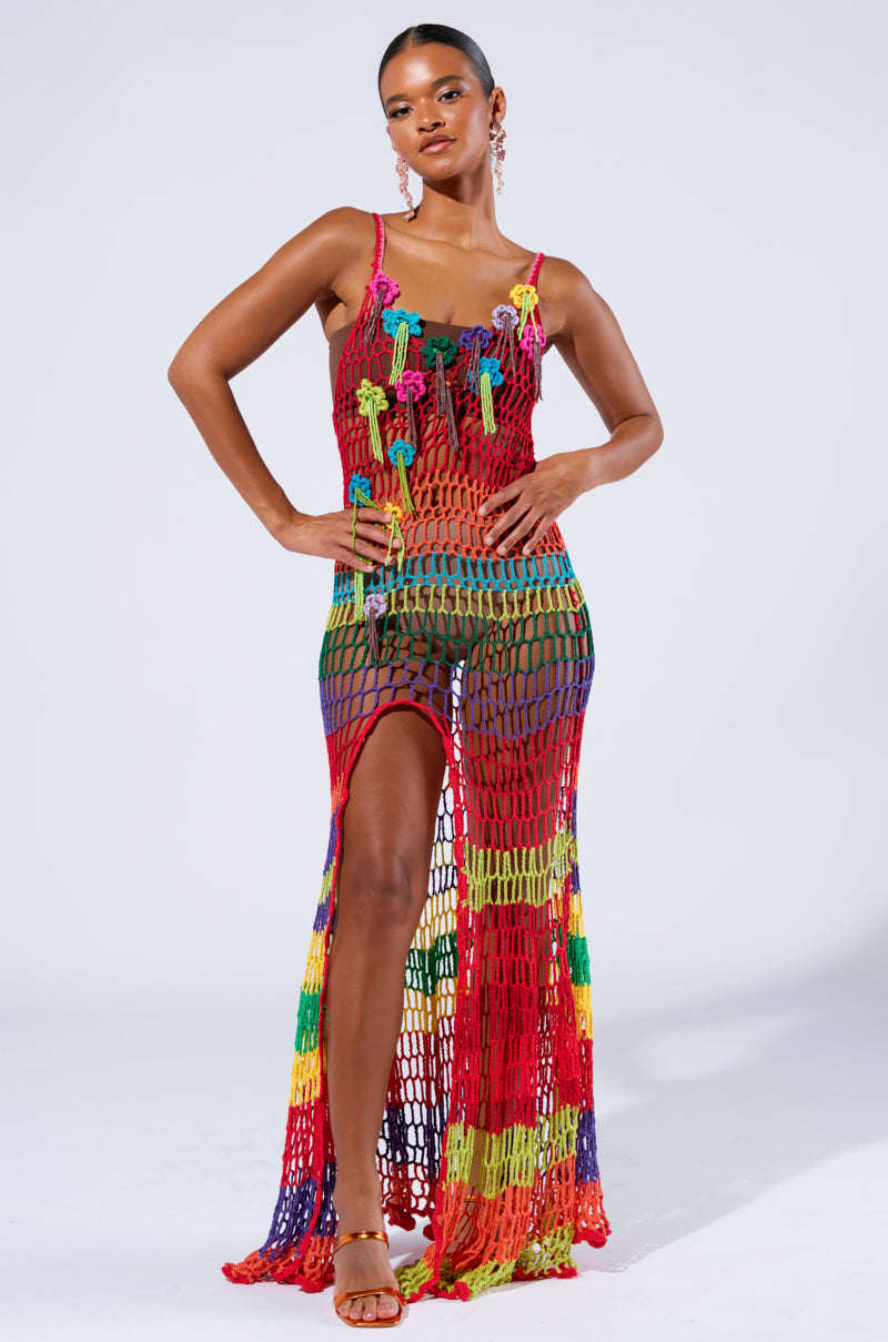 A woman poses confidently against a plain white background, dressed in the eye-catching OCEAN BREEZE CROCHET MAXI DRESS. The vibrant hues of red, green, yellow, and purple mix beautifully on this high-slit beach-ready dress. She pairs it perfectly with strappy sandals and hoop earrings, her hair elegantly pulled back.
