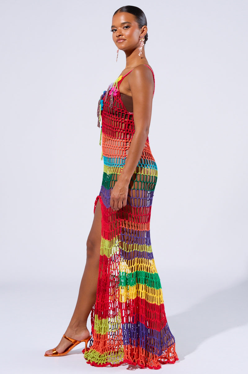 A woman is striking a pose in the OCEAN BREEZE CROCHET MAXI DRESS, showcasing its rainbow-striped crochet-knit fabric and high side slit. She completes her stylish ensemble with orange high-heeled sandals and statement earrings, her hair elegantly pulled back against a plain white background.