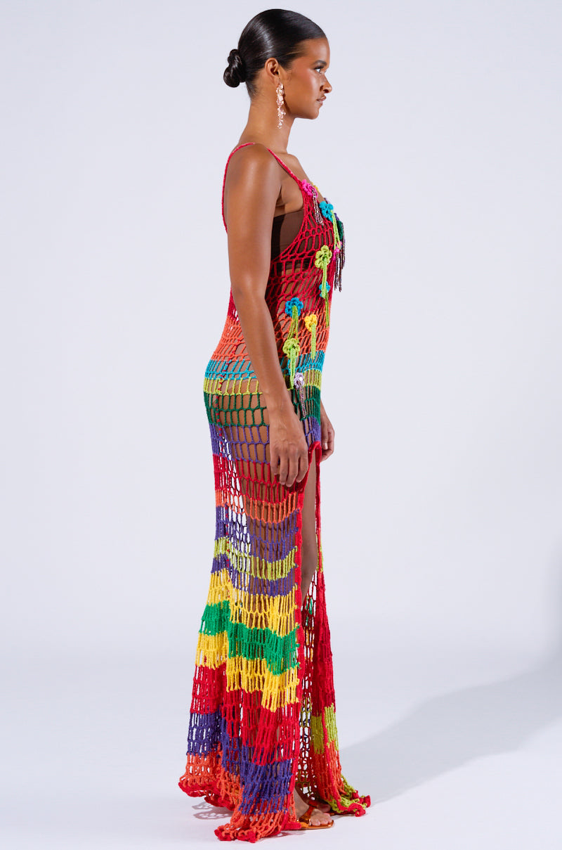 A woman stands in profile against a plain background, wearing the OCEAN BREEZE CROCHET MAXI DRESS, a long, colorful, and vibrant rainbow-patterned crochet maxi dress. The semi-transparent fabric reveals a black garment underneath. With her hair pulled back and earrings sparkling, she exudes confidence in this beach-ready ensemble.