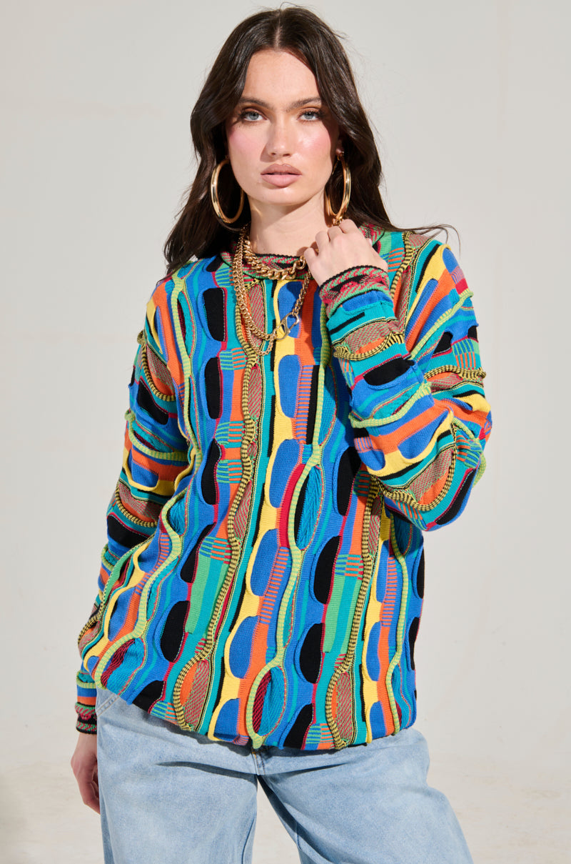 A person with long dark hair poses confidently, wearing the VINTAGE COOL OVERSIZED MULTI COLOR SWEATER paired with blue jeans. They have large hoop earrings and hold the sweater collar with one hand against a plain, light-colored background.