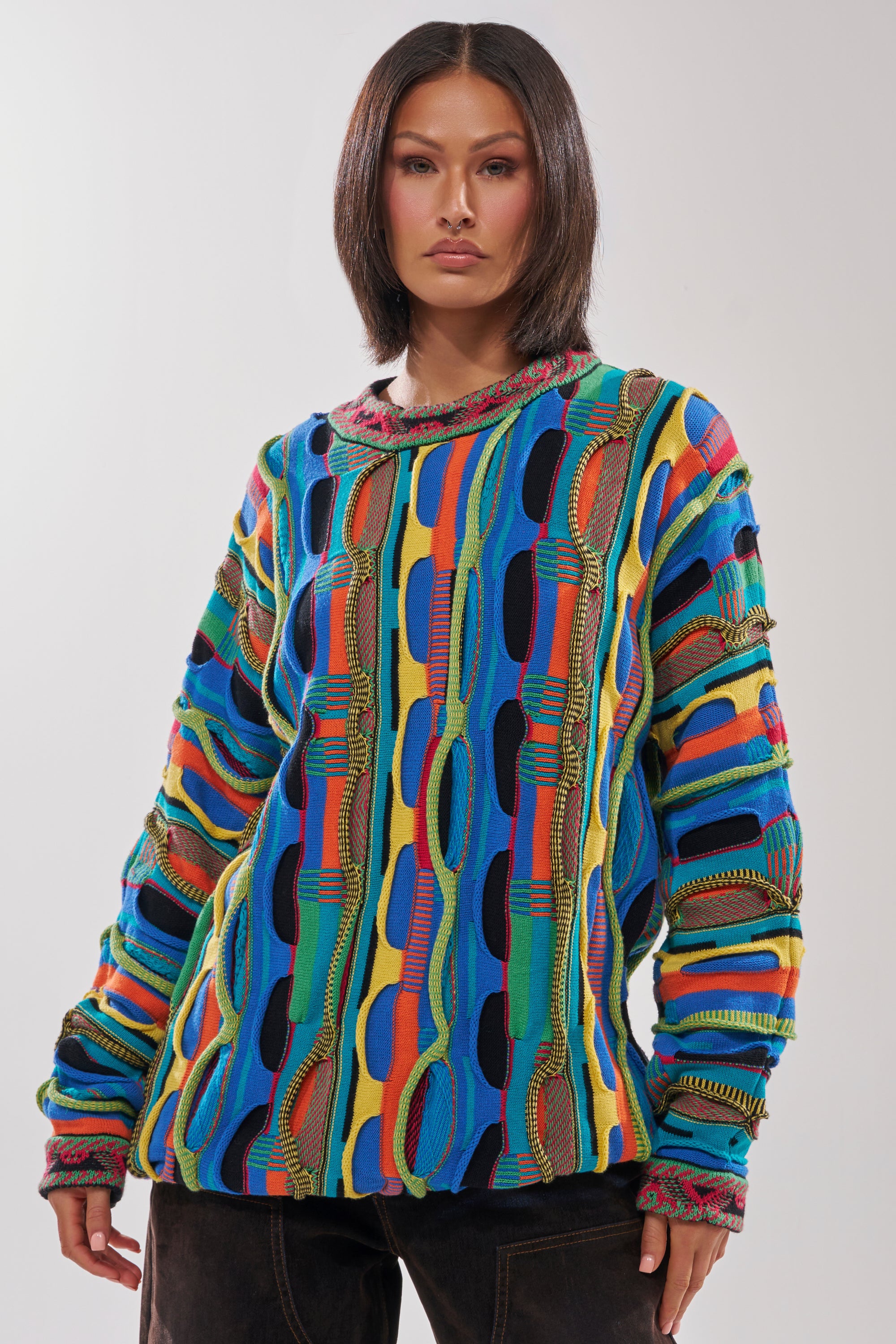 A person with straight, shoulder-length dark hair models the VINTAGE COOL OVERSIZED MULTI COLOR SWEATER, featuring abstract blue, yellow, red, and green patterns, standing against a plain light background.