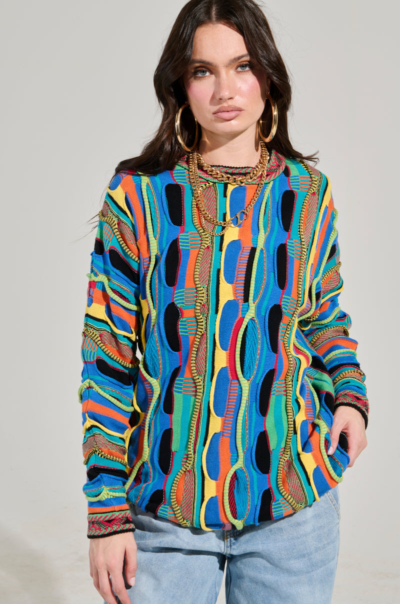 A person with long dark hair stands confidently, wearing the VINTAGE COOL OVERSIZED MULTI COLOR SWEATER, featuring vibrant retro-inspired knit designs in blue, yellow, red, and green. To complement this statement piece, they pair light blue jeans and large hoop earrings while posing against a plain background.