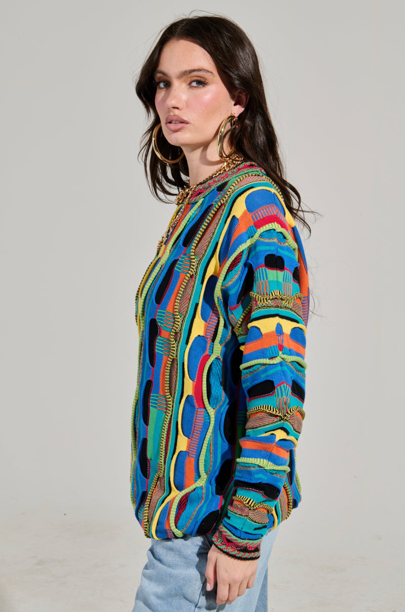 A person with long, dark hair models the VINTAGE COOL OVERSIZED MULTI COLOR SWEATER, showcasing its retro-inspired wavy lines and geometric shapes. They accessorize with large hoop earrings and pair it with light blue jeans, standing against a plain gray background.