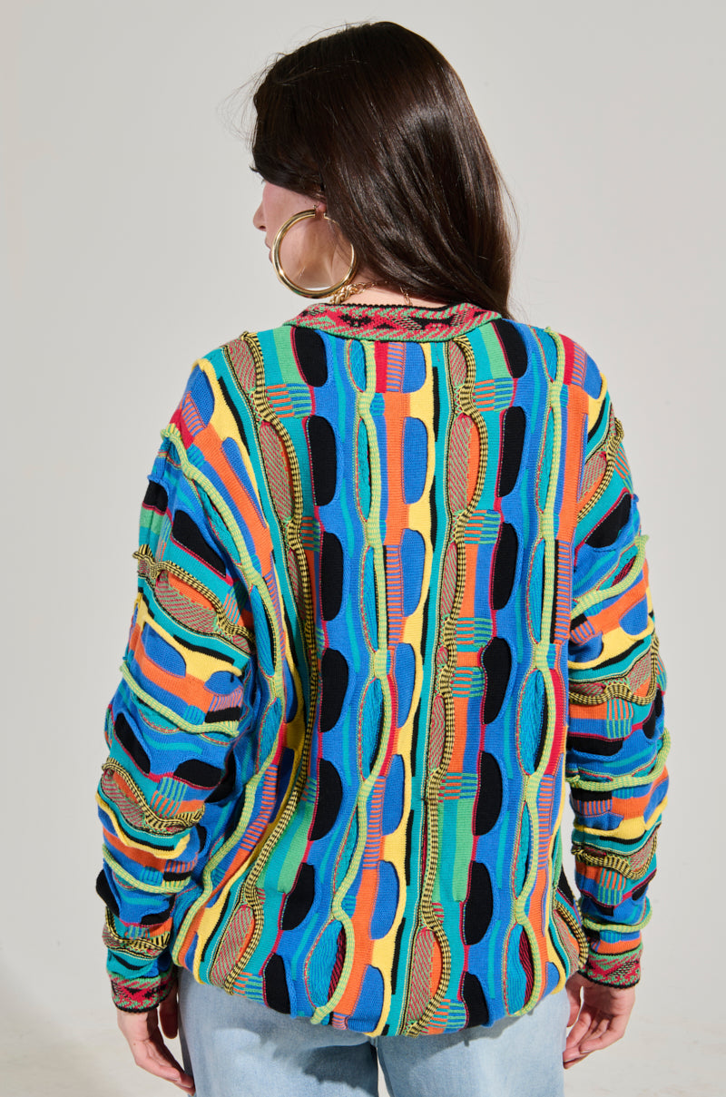 A person with long dark hair, facing away, is wearing the VINTAGE COOL OVERSIZED MULTI COLOR SWEATER, featuring bold wavy patterns in blue, yellow, red, and green. This sweater pairs perfectly with large hoop earrings and light blue jeans.