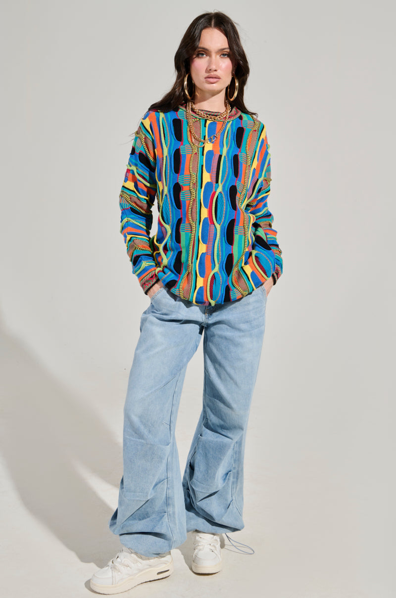 A person with long hair stands against a plain background, showcasing the VINTAGE COOL OVERSIZED MULTI COLOR SWEATER paired with light blue wide-legged jeans. They complete the look with white sneakers and layered necklaces.