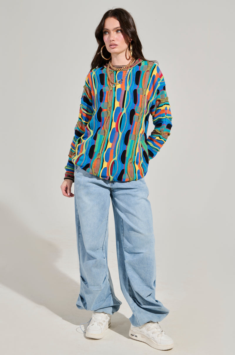 A person confidently stands against a neutral background, wearing the VINTAGE COOL OVERSIZED MULTI COLOR SWEATER, paired with baggy light blue jeans and white sneakers. They have long hair adorned with large hoop earrings and layered necklaces.
