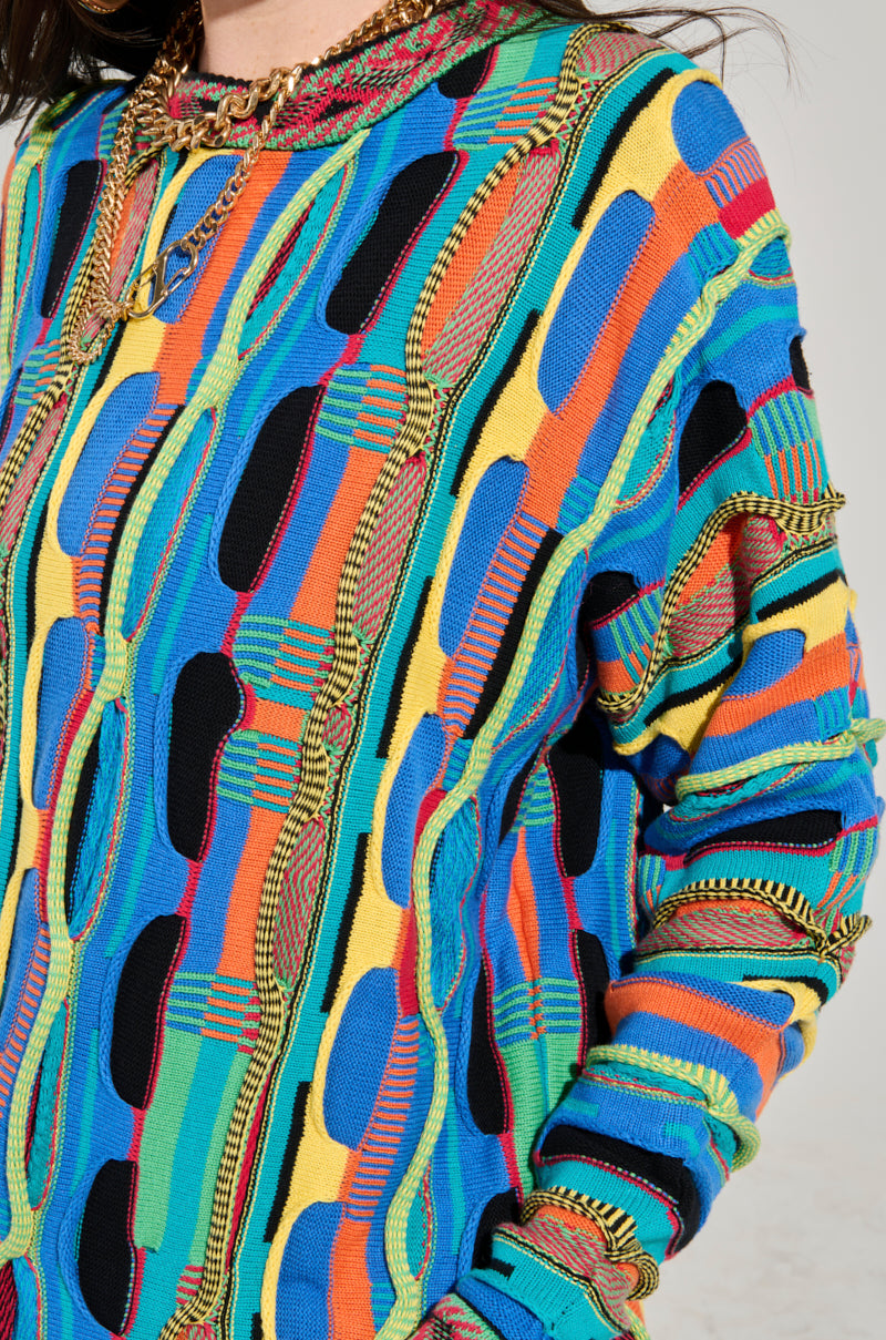 Close-up of someone wearing the VINTAGE COOL OVERSIZED MULTI COLOR SWEATER, featuring a retro-inspired knit design with abstract patterns in blue, orange, yellow, and green. This unique sweater highlights an array of intricate woven shapes and lines.