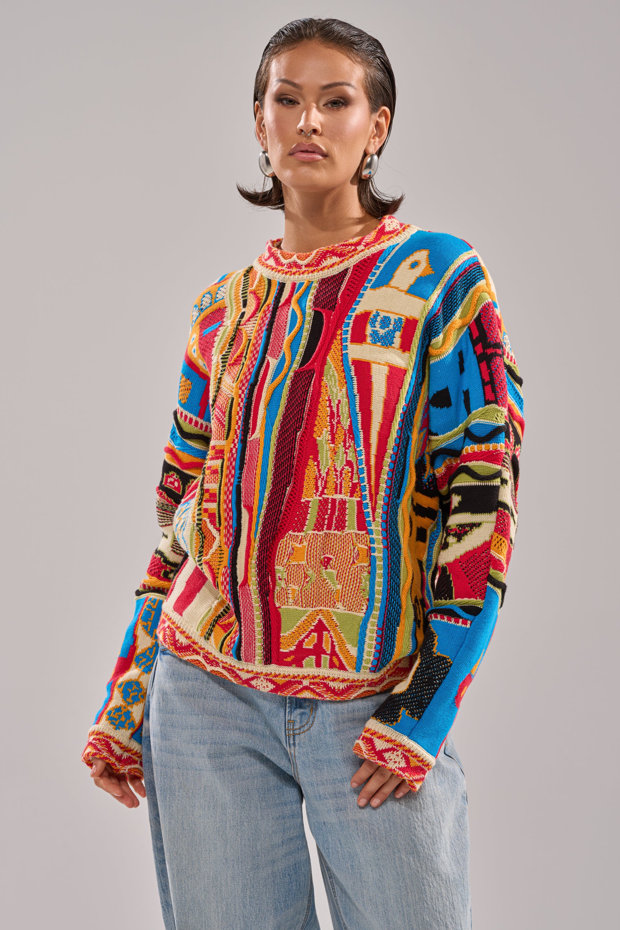 A woman in the RETRO OVERSIZED SWEATER IN MULTI, featuring abstract red, blue, yellow, and black patterns, pairs it with light blue jeans and stands confidently against a plain gray background.