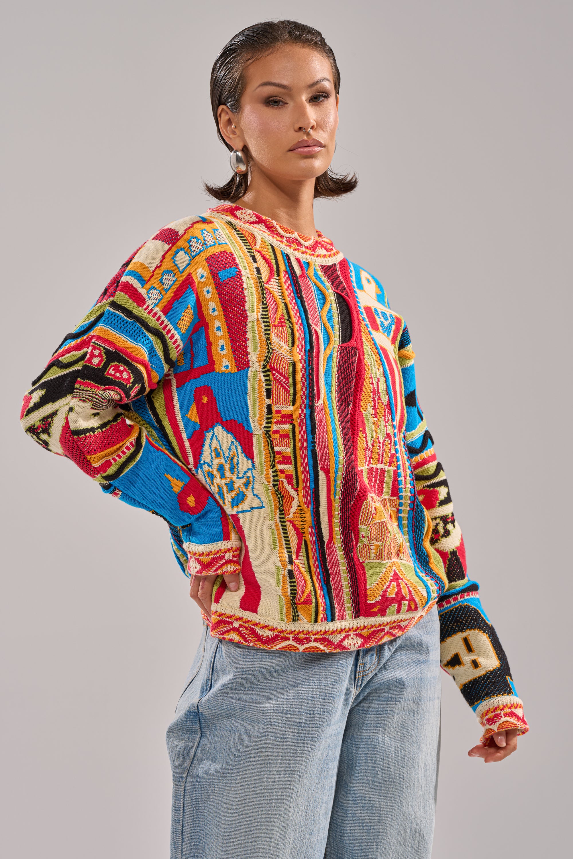 A woman with slicked-back hair models the RETRO OVERSIZED SWEATER IN MULTI—featuring bold red, blue, yellow, and cream tones—with light blue jeans, standing confidently against a plain light background.