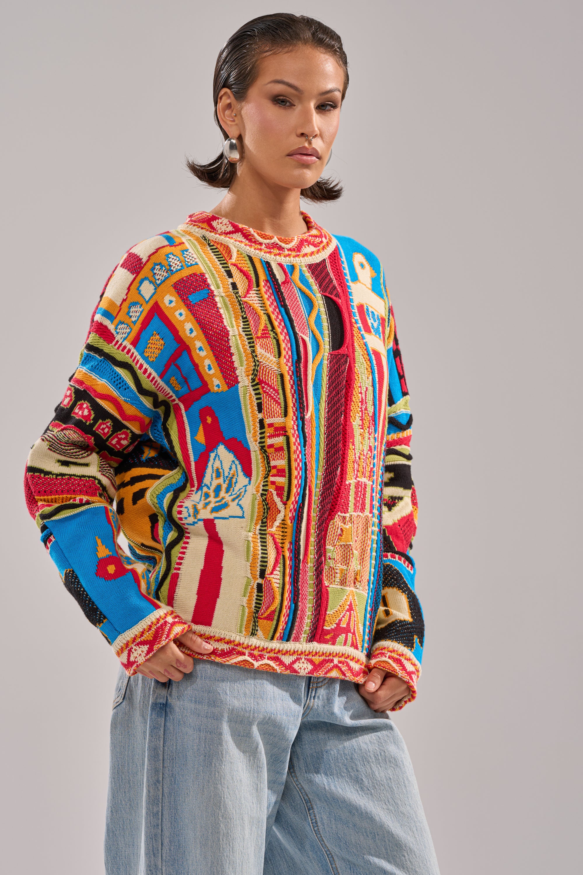 A person models the RETRO OVERSIZED SWEATER IN MULTI, featuring colorful, abstract patterns. They pair it with light blue jeans and have short, slicked-back hair, standing before a plain light gray background.