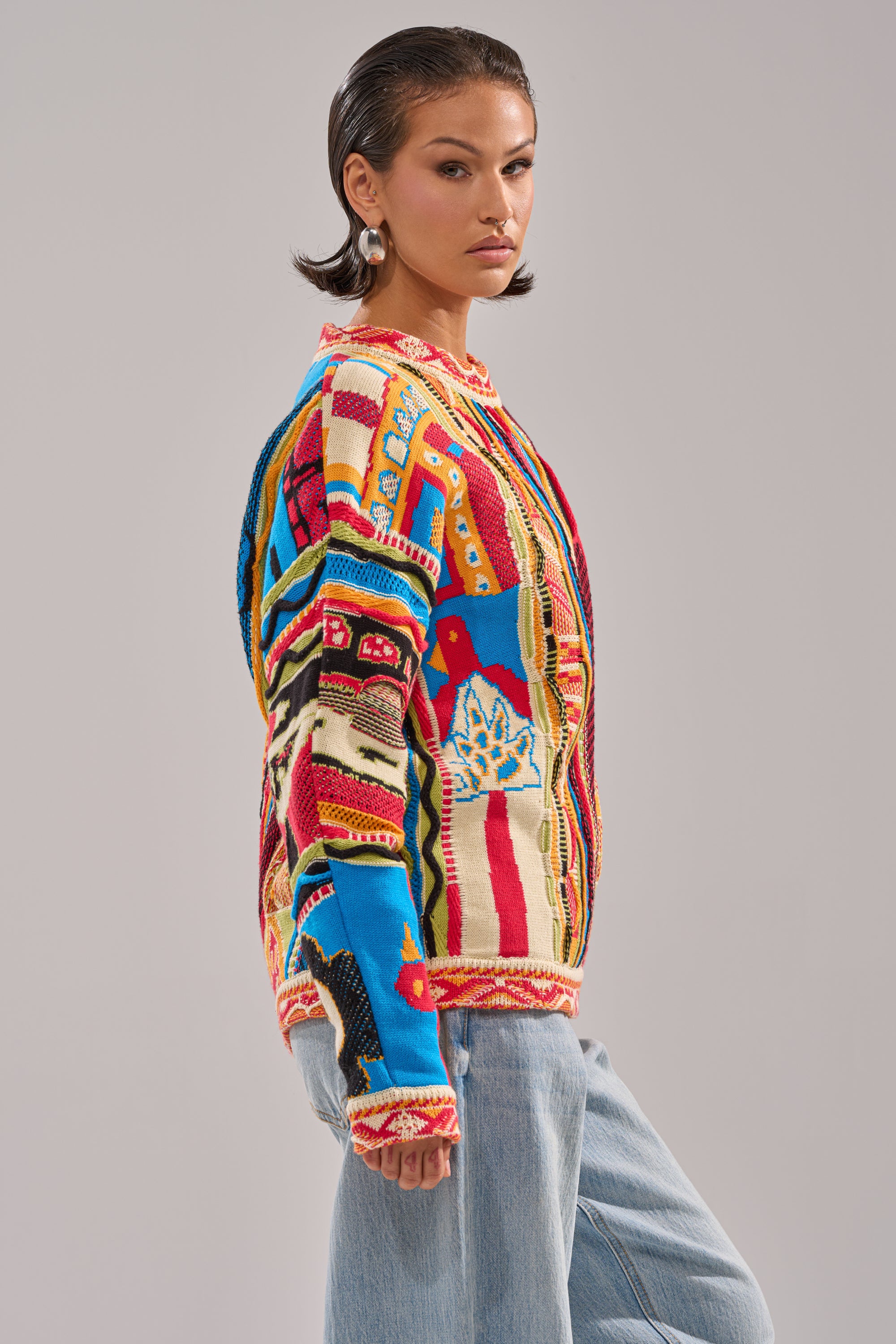 A woman with short brown hair and hoop earrings wears the RETRO OVERSIZED SWEATER IN MULTI and light blue jeans, standing confidently against a plain gray background.