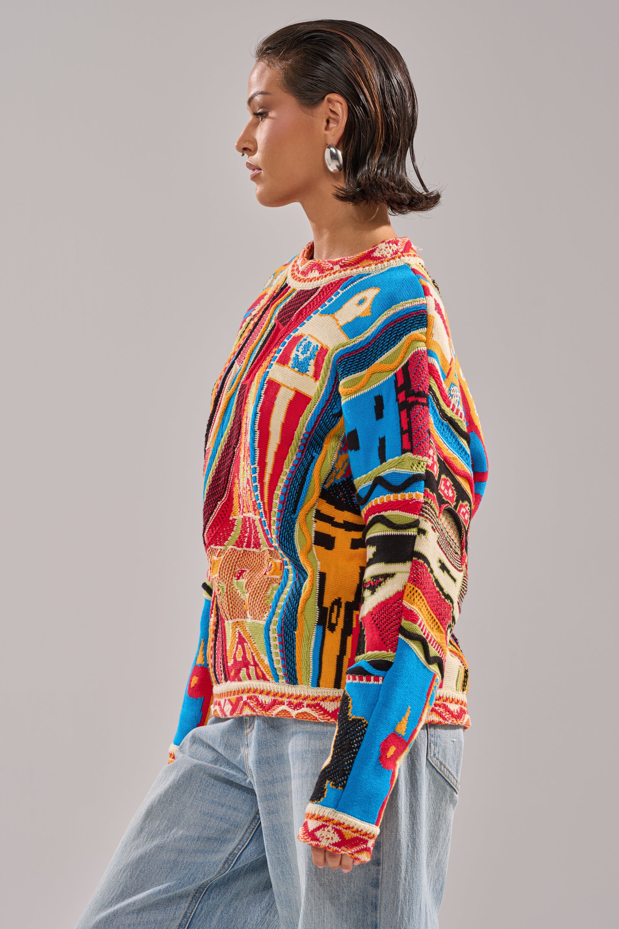 A person with short, slicked-back hair models the RETRO OVERSIZED SWEATER IN MULTI with light blue jeans, standing in profile against a plain gray background.